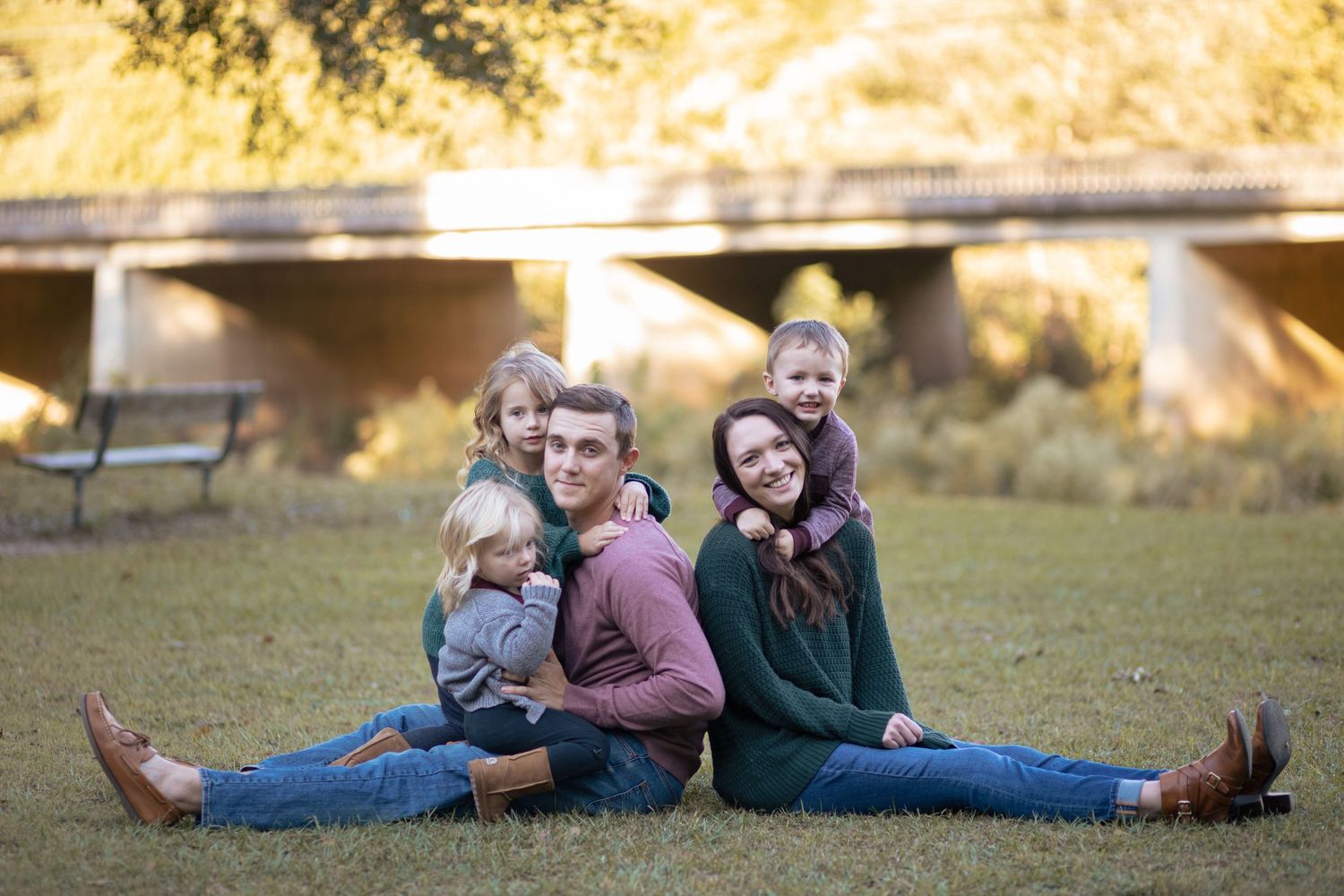 Family of five sitting in grass, parents back-to-back with three young children, bridge in background.