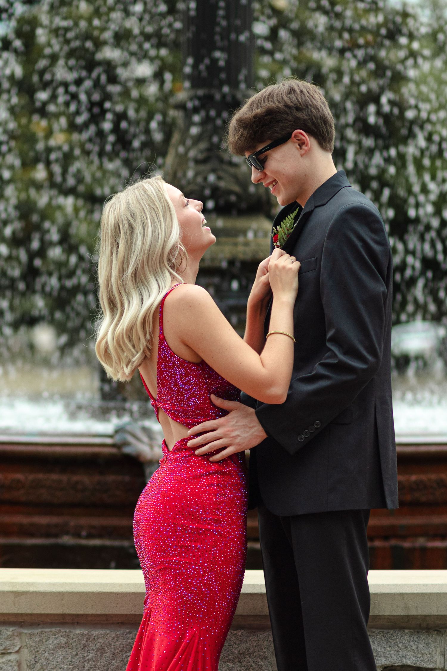 Couple dressed in prom attire embrace in front of a fountain outdoors.