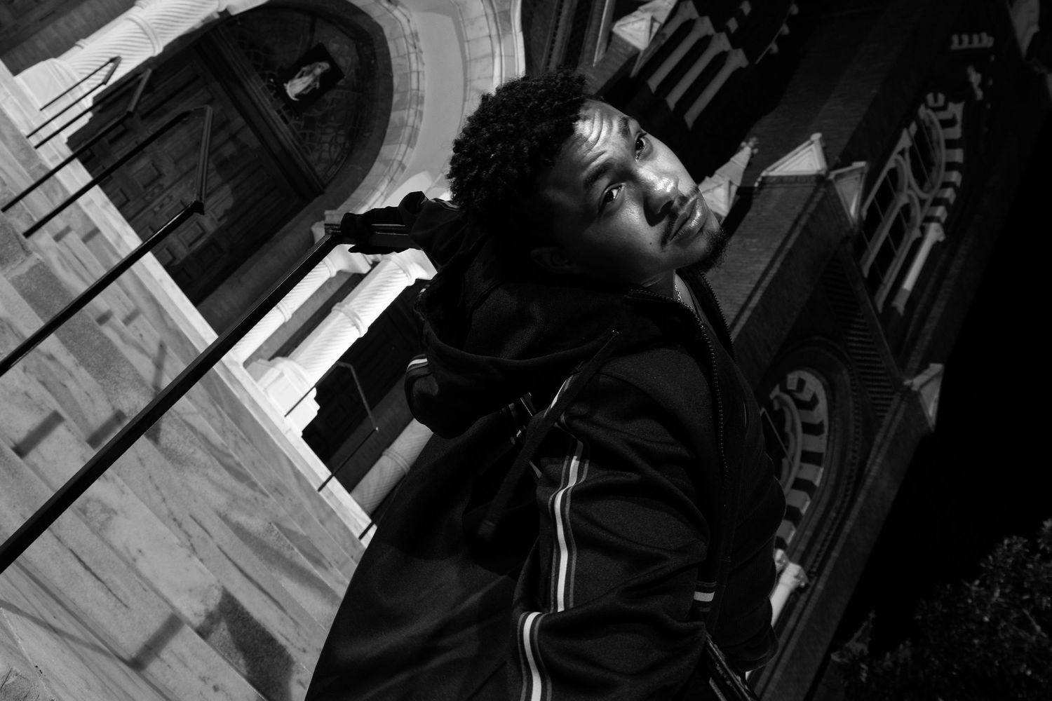 Black and white photo of a young man in a black jacket posing against a historic stone church building.