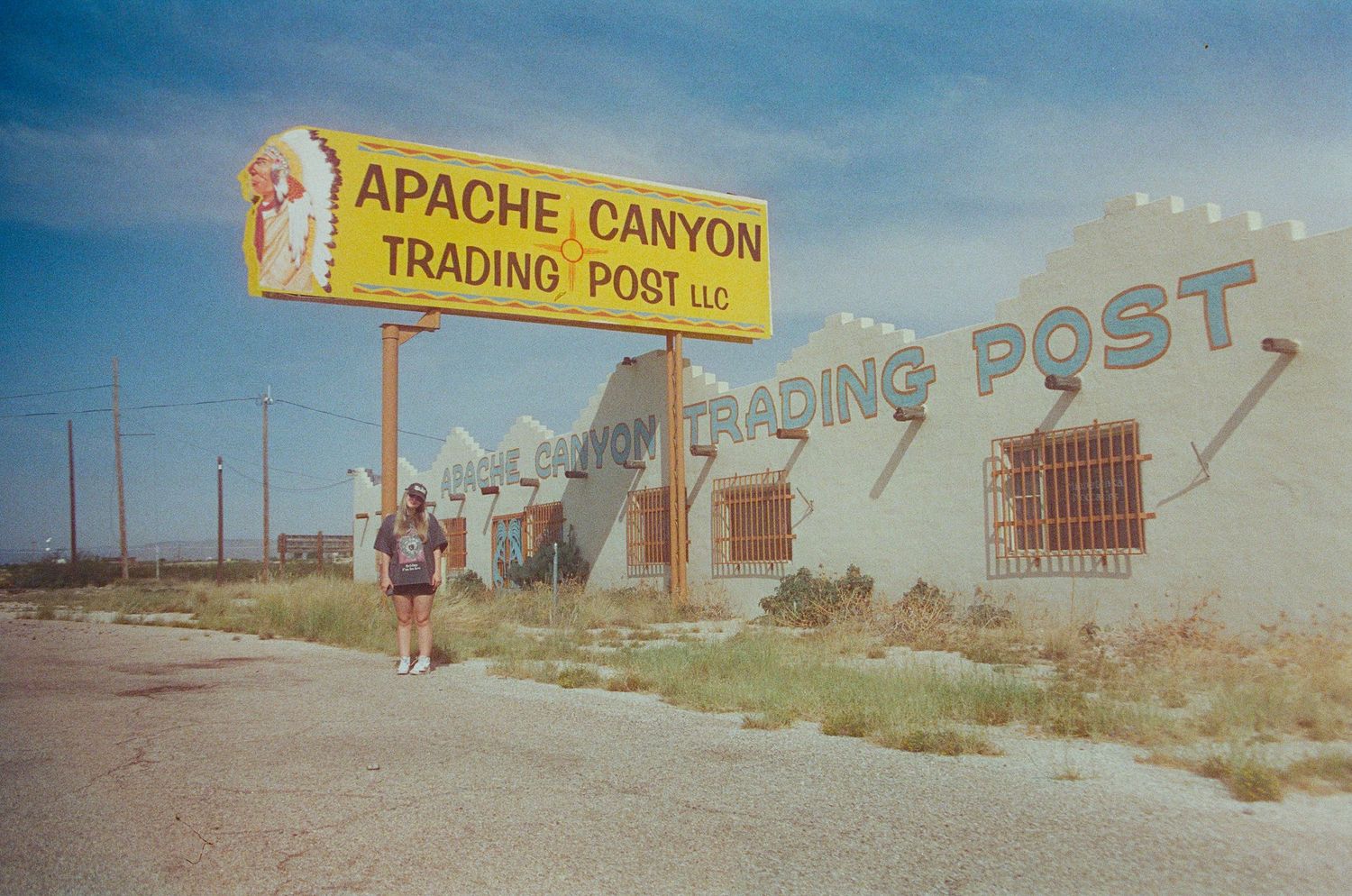 A person stands beside the Apache Canyon Trading Post LLC sign in a desert Southwest location.
