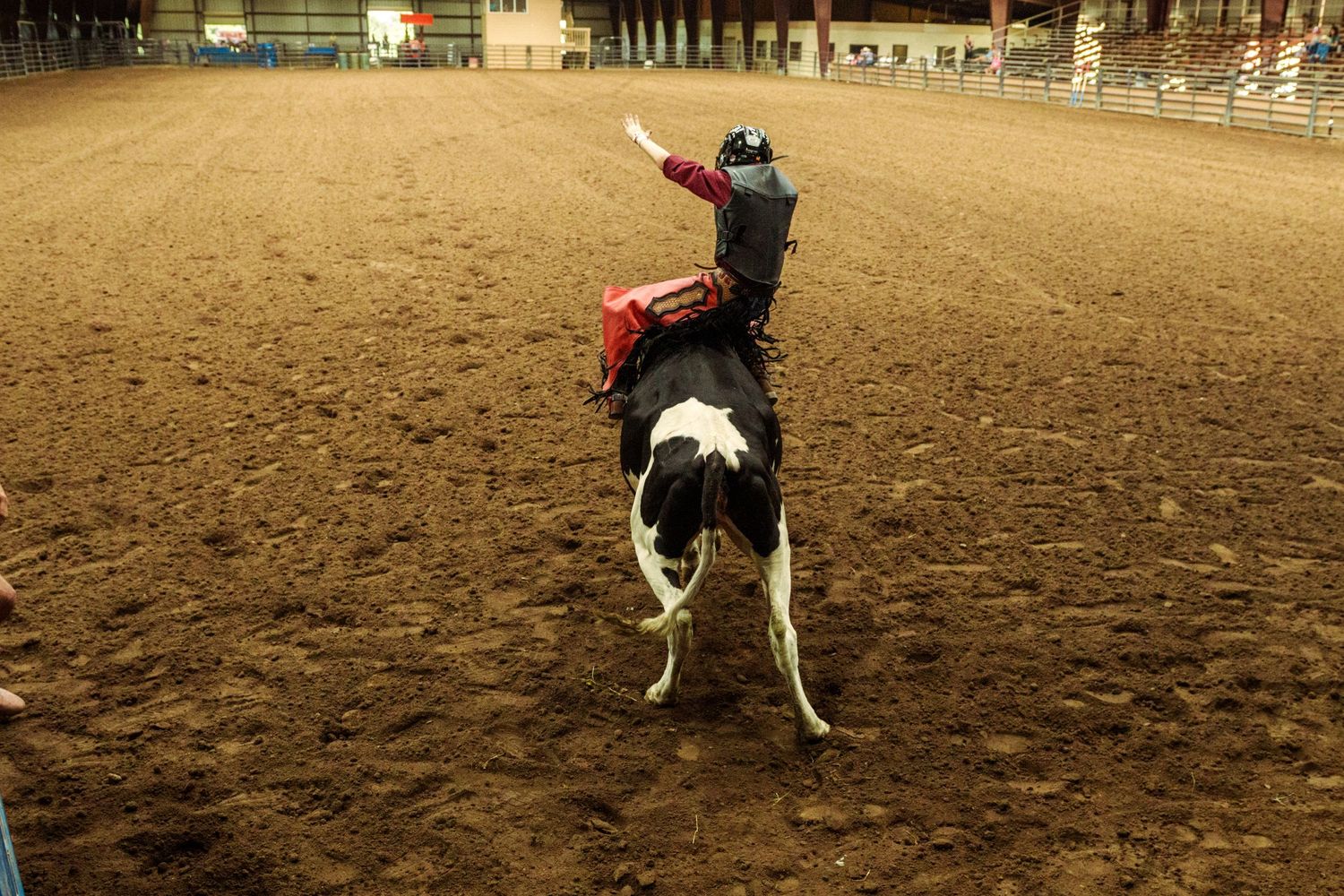 A rodeo rider on a black and white bull during an indoor rodeo event on a dirt arena floor.