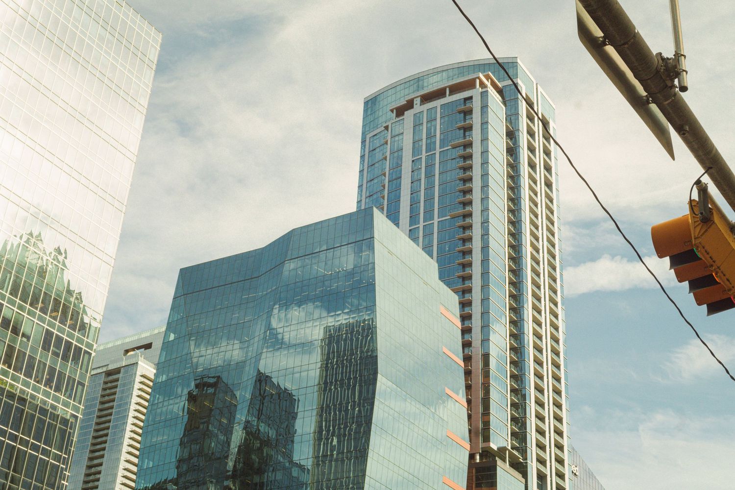 Modern glass skyscrapers reflect clouds in a downtown cityscape with traffic lights visible against a partly cloudy sky.