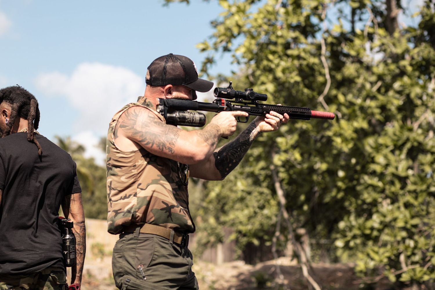 Tattooed man in tactical vest aiming rifle with scope outdoors at shooting range.