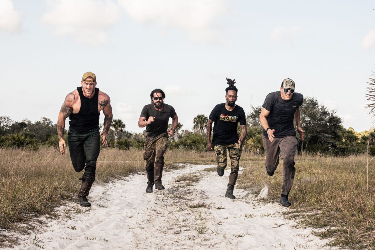 Four athletic men running on a sandy trail through grassy fields in black and camo workout gear.