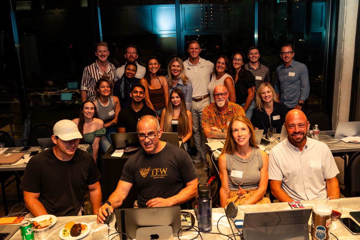 A large group of smiling professionals poses together in a modern office setting during a team event at night.