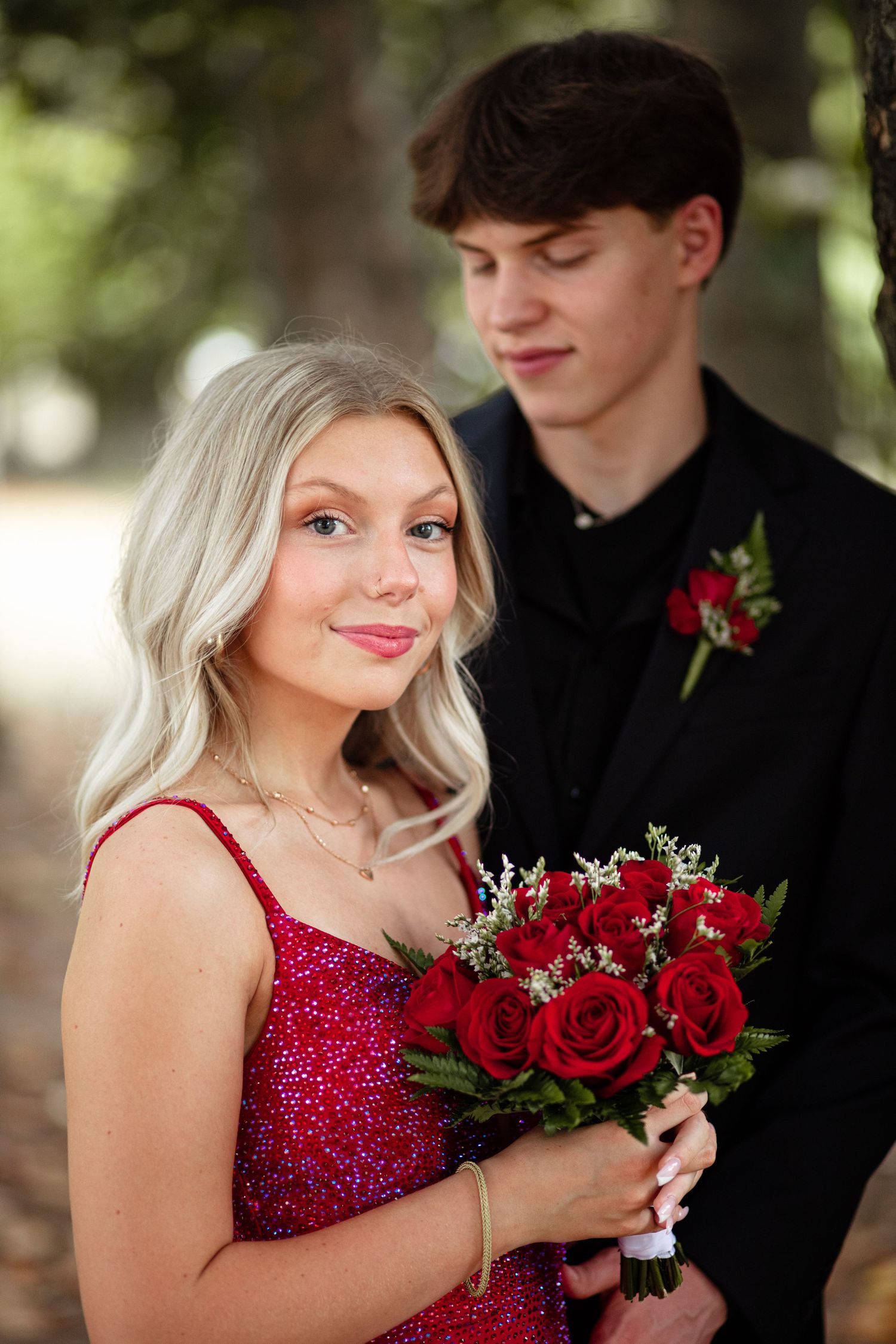 Teen prom couple posing outdoors, girl in red sequin dress holding red roses with boy in black suit.