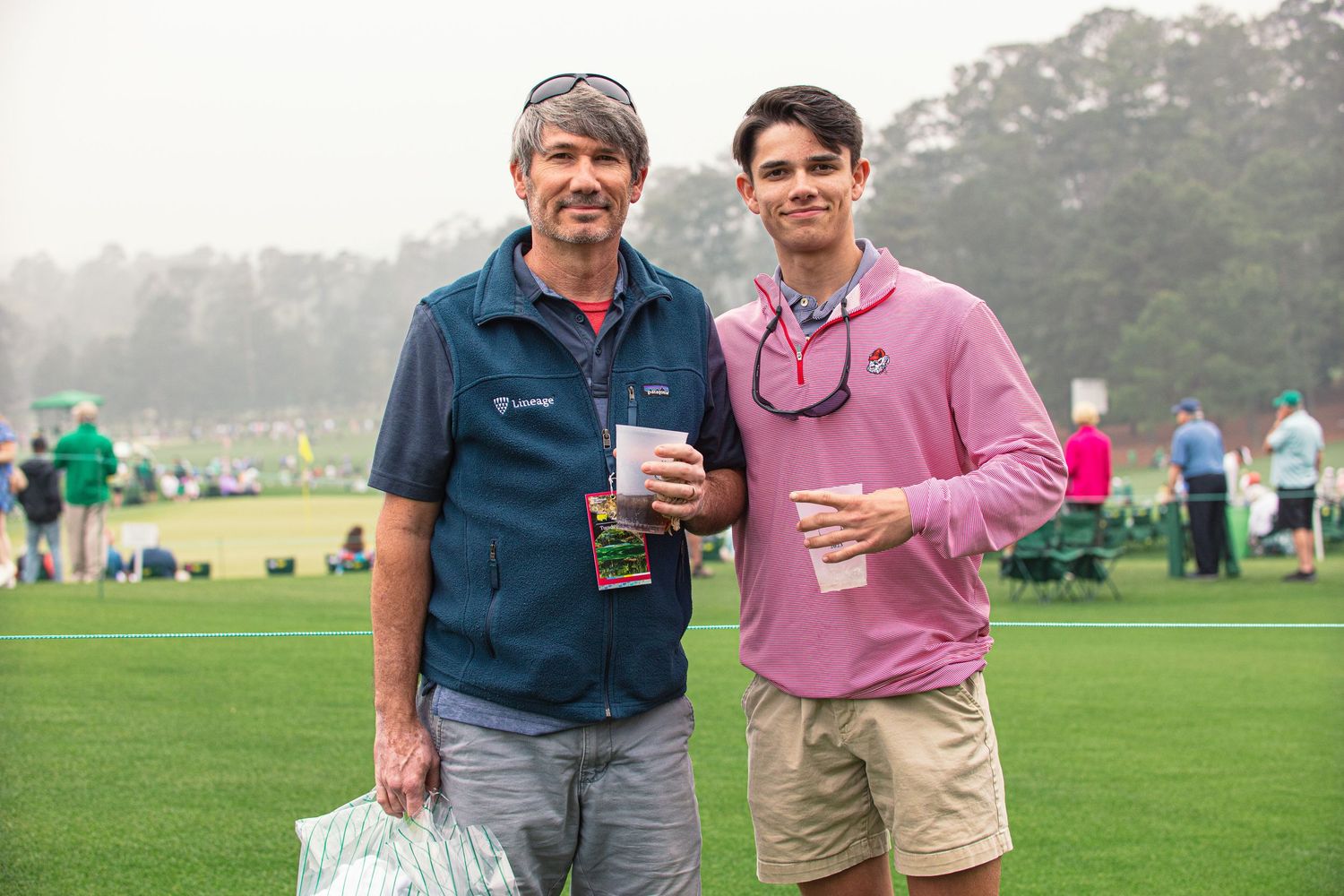 Two men pose together on a golf course, one in a blue vest and one in a pink shirt, holding drinks at an outdoor event.