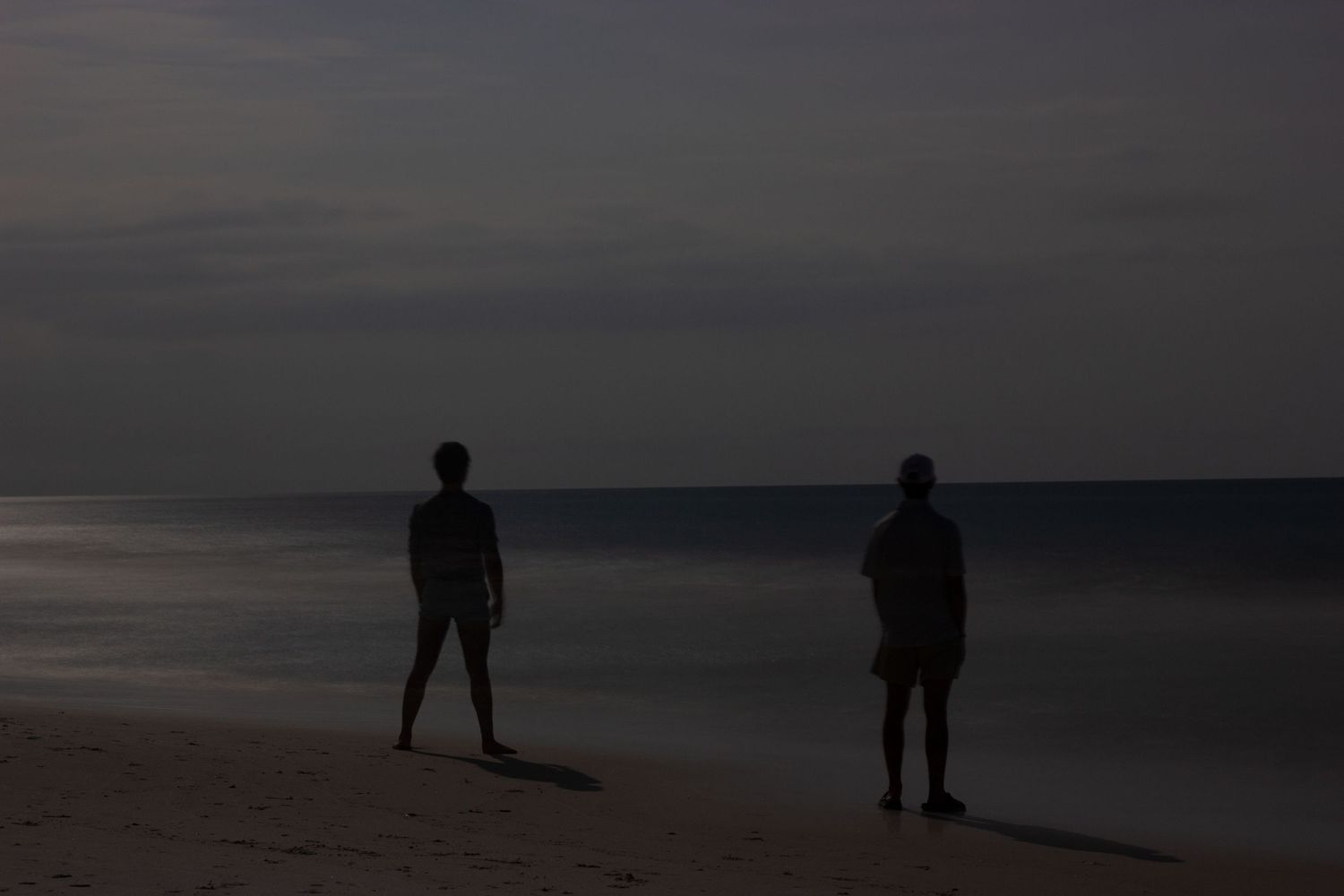 Two silhouettes stand on a dark beach at dusk, with calm ocean waves and overcast sky in the background.