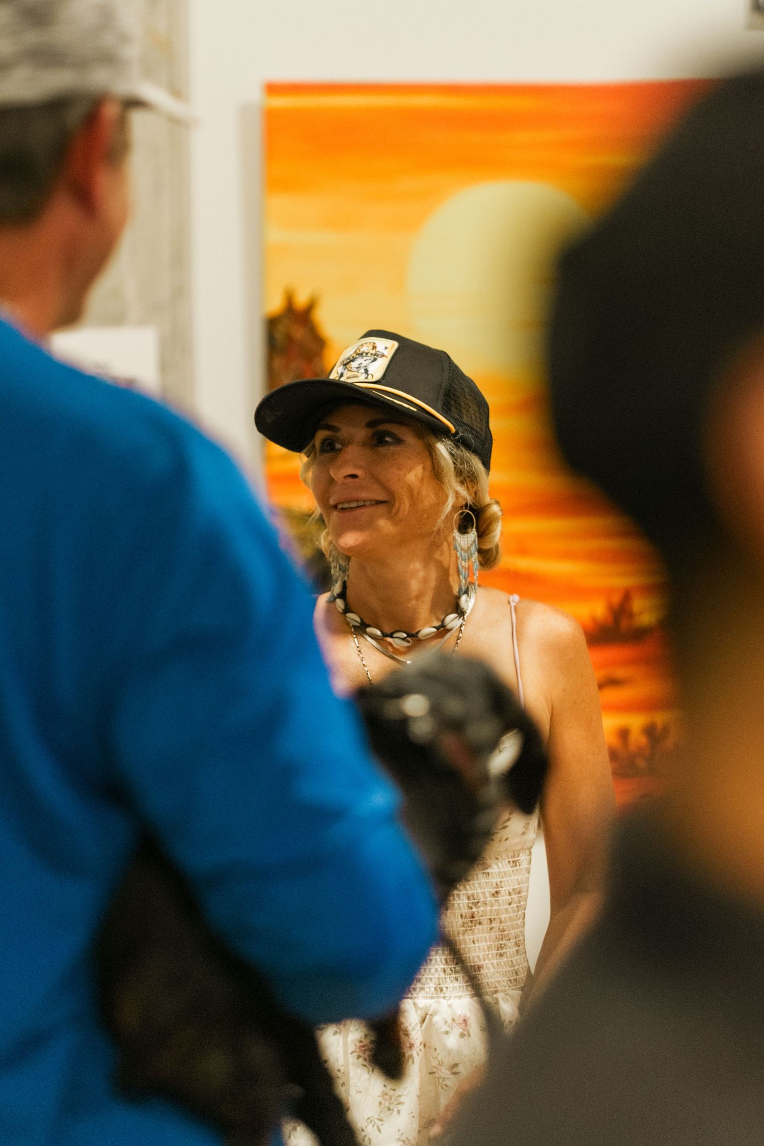 Woman in black cap and floral dress smiling at an art gallery event with colorful paintings in background.