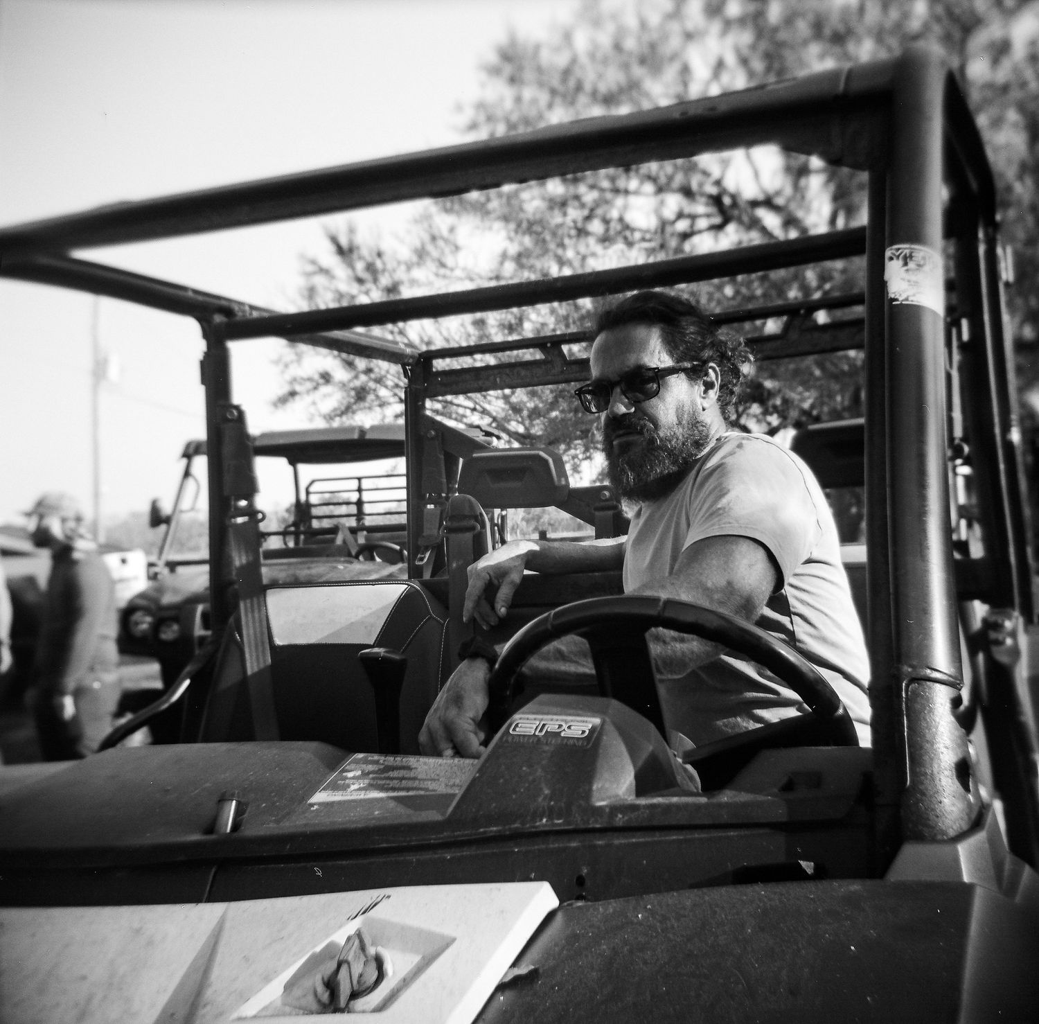 Bearded man in sunglasses sits behind the wheel of an open-top vehicle in black and white photo.