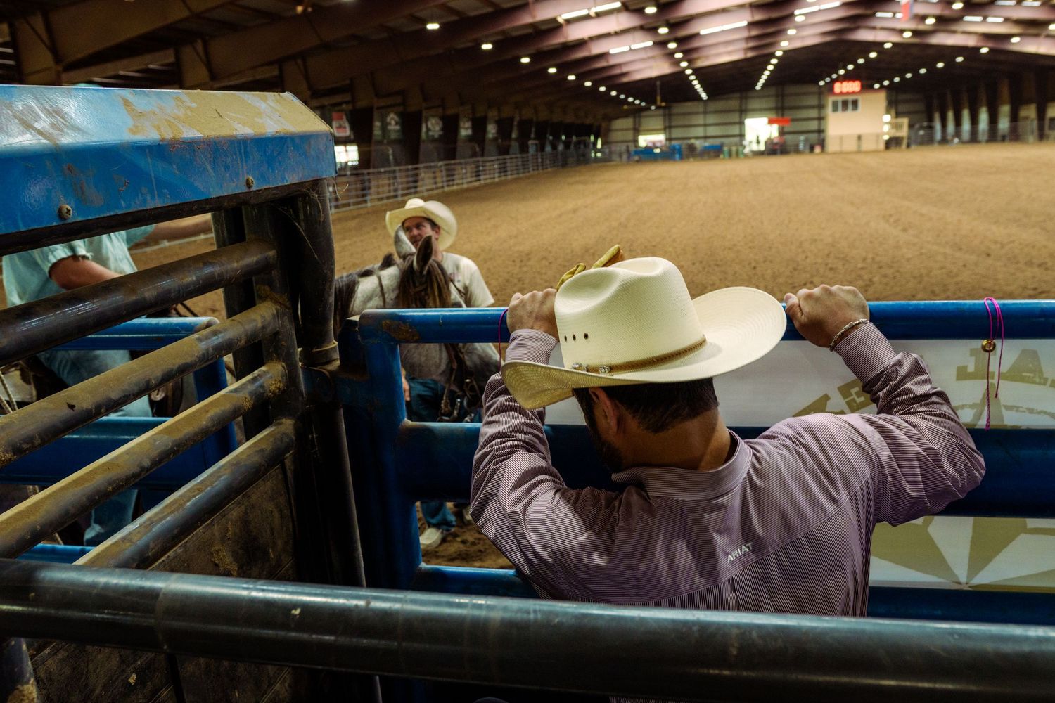 Cowboys in western hats prepare behind the chutes at an indoor rodeo arena, ready for competition.