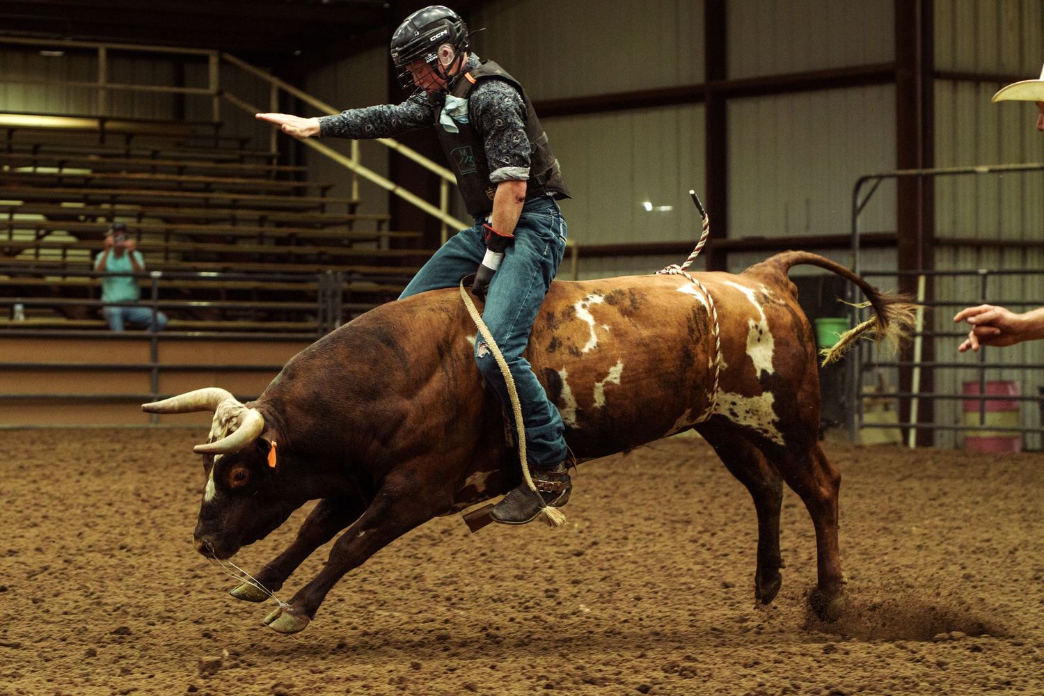 A bull rider grips a bucking brown bull with horns during an indoor rodeo competition on a dirt arena floor.