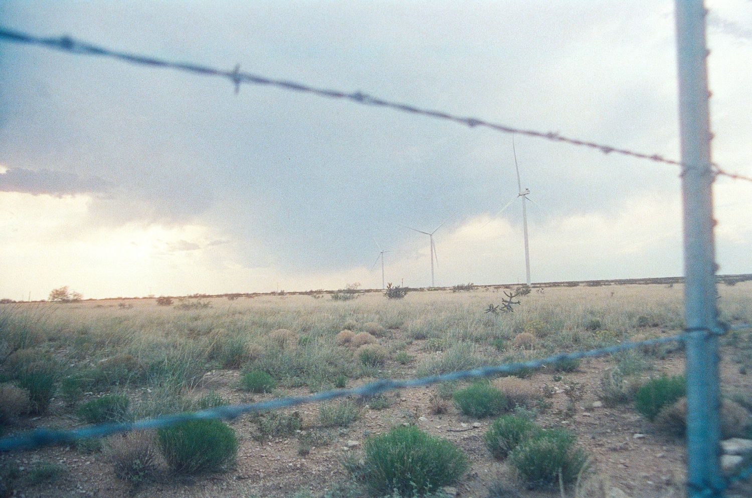 Barbed wire fence in arid desert landscape with sparse shrubs, communication towers, and stormy sky in background.
