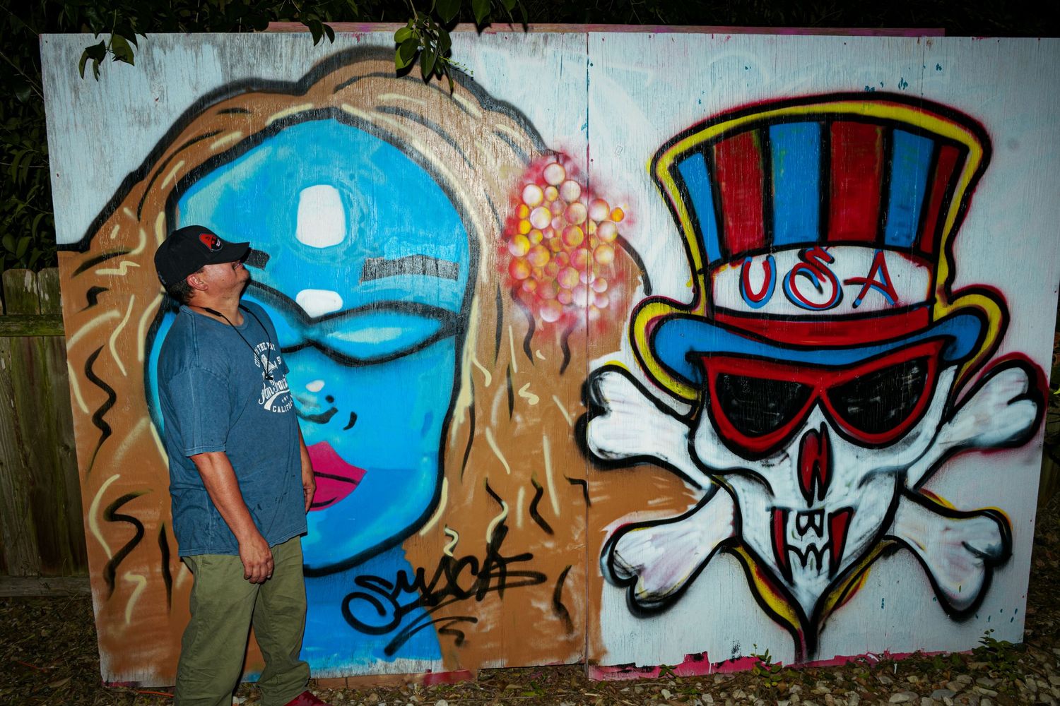 Man posing beside colorful graffiti mural featuring blue face and USA skull with top hat on wooden wall.