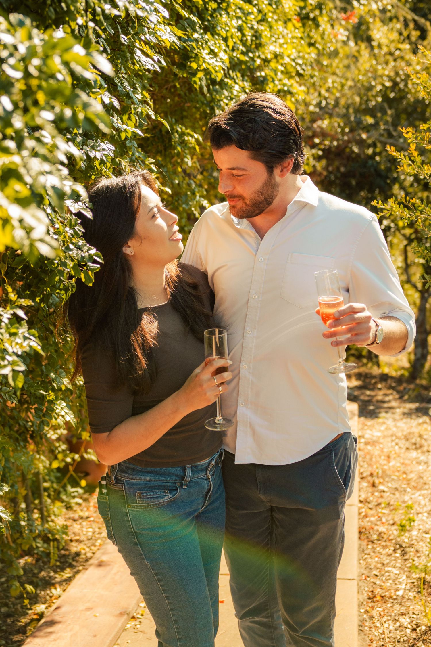 Couple sharing a romantic moment with wine glasses in a sunny vineyard garden, surrounded by lush green foliage.