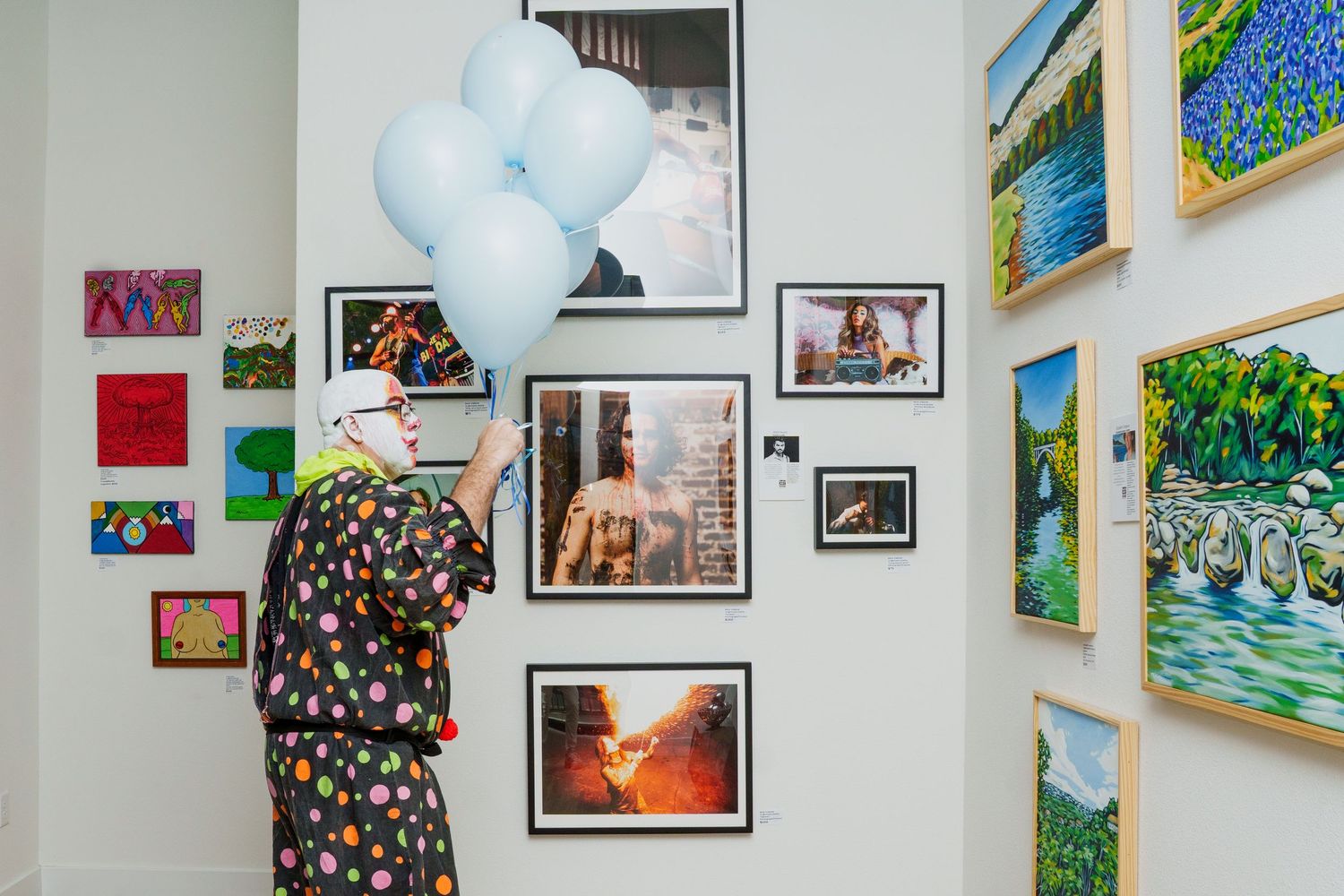 A clown in colorful polka-dot costume holds light blue balloons in an art gallery with framed paintings on white walls.