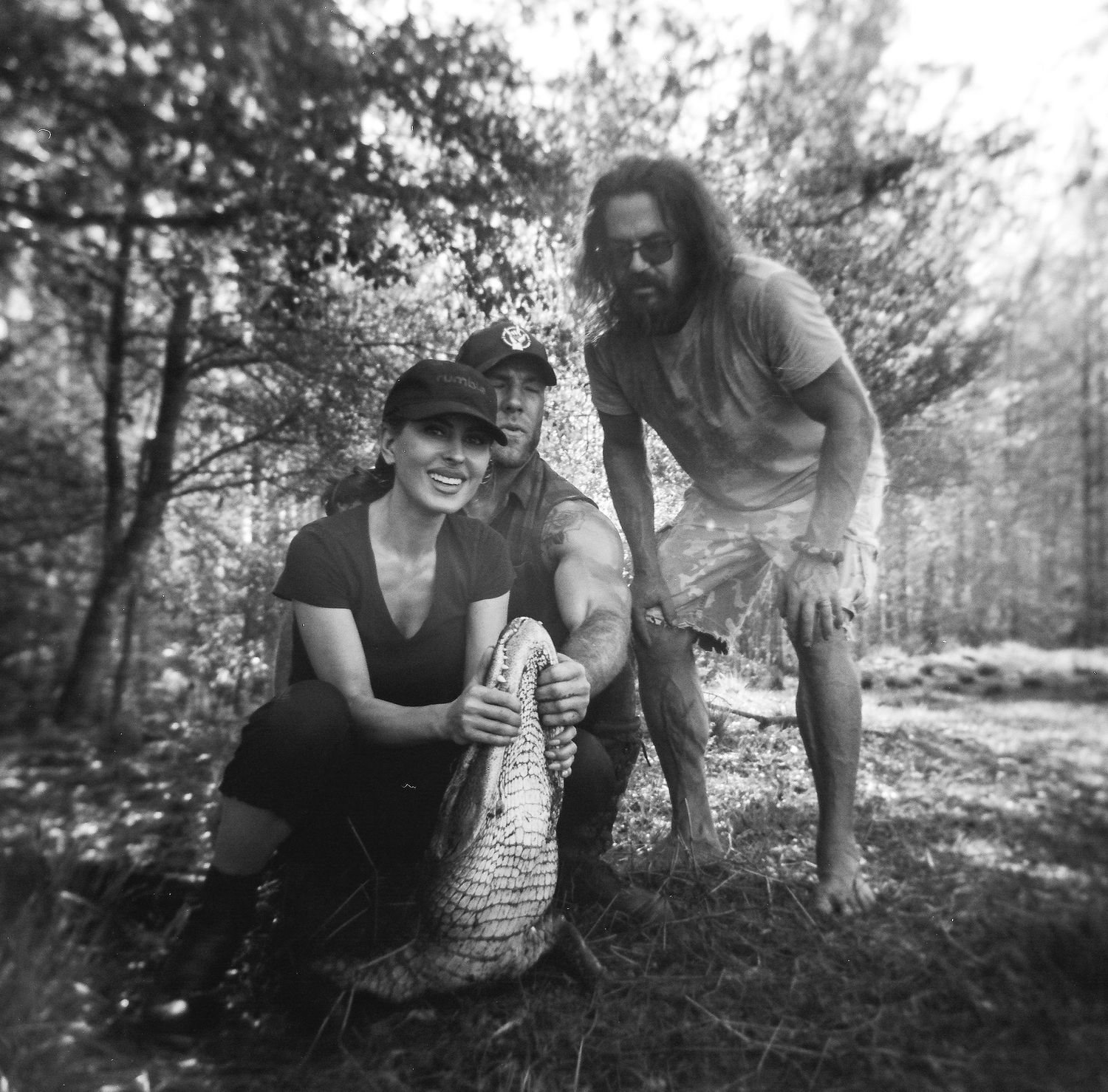 Black and white photo of two people posing with a large alligator in a wooded outdoor setting.
