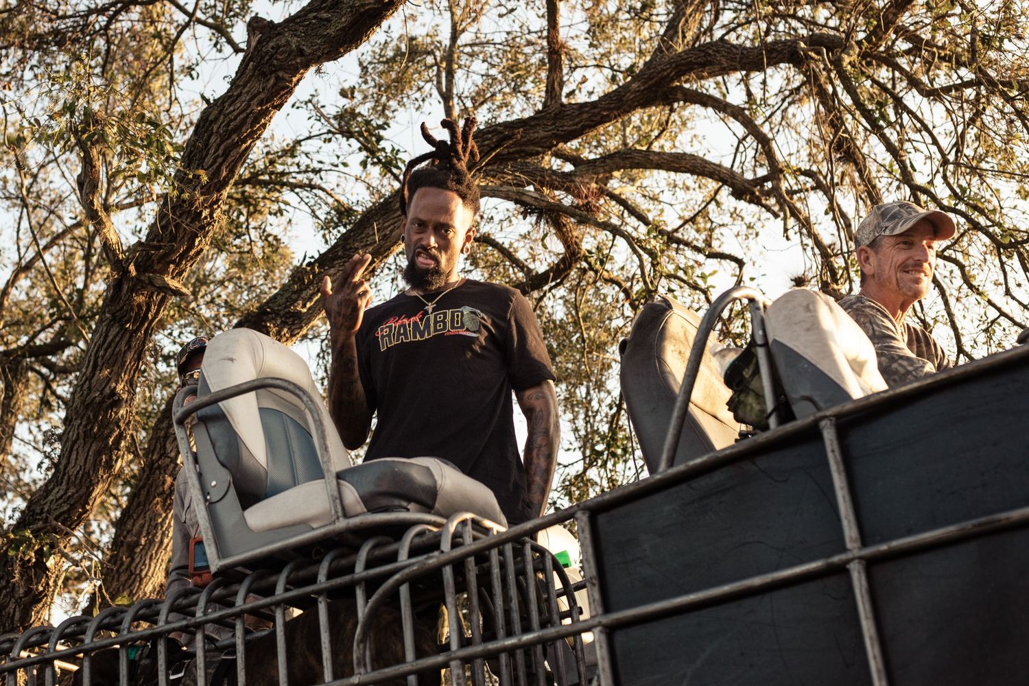 Two men standing on a flatbed truck outdoors, surrounded by large oak trees in natural daylight.