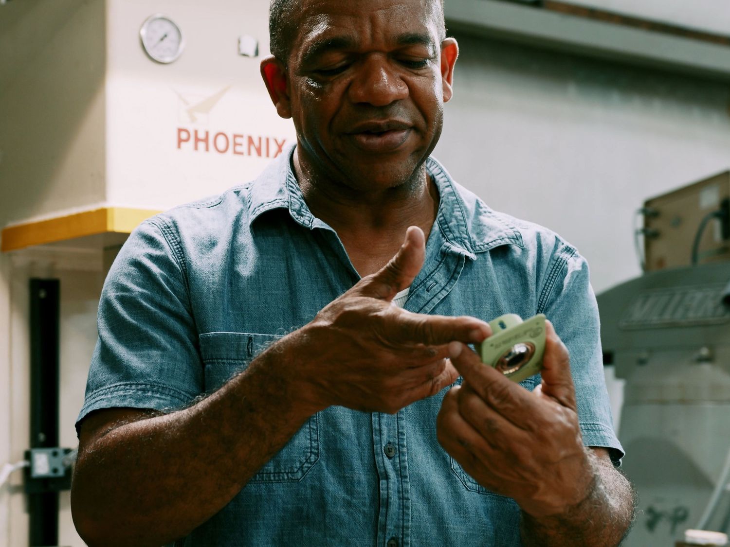 Experienced machinist examines a small green metal part near a Phoenix machine.