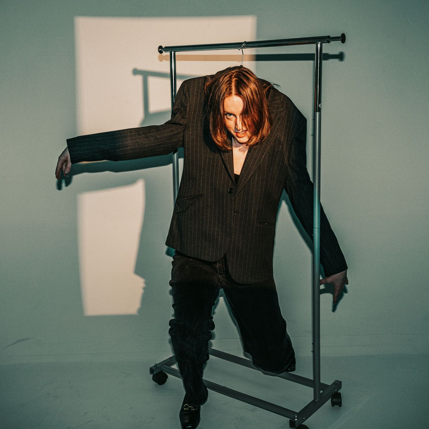 Person posing through metal clothing rack wearing oversized pinstripe blazer in studio setting.