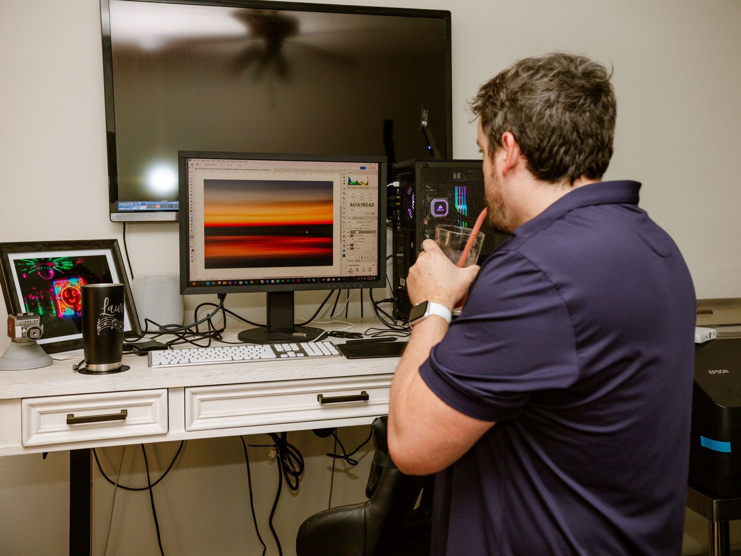 Man in navy polo calibrating monitors displaying a sunset landscape photo at a desk.