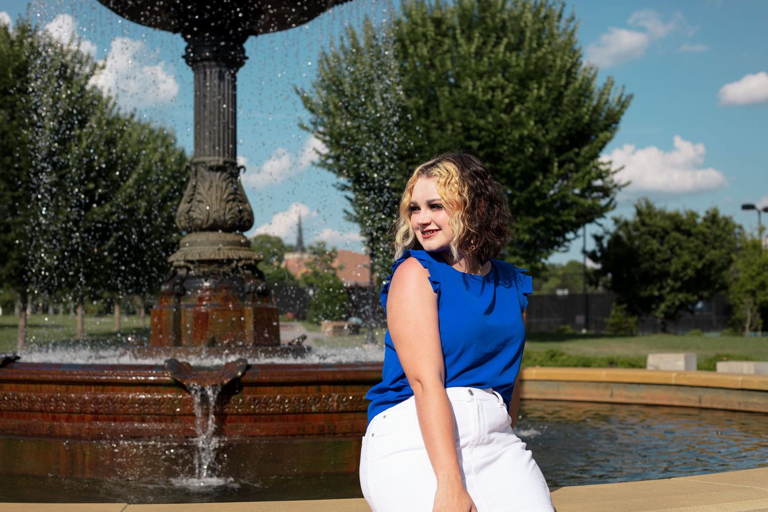 Blonde woman in blue top and white pants posing near an ornate outdoor fountain.