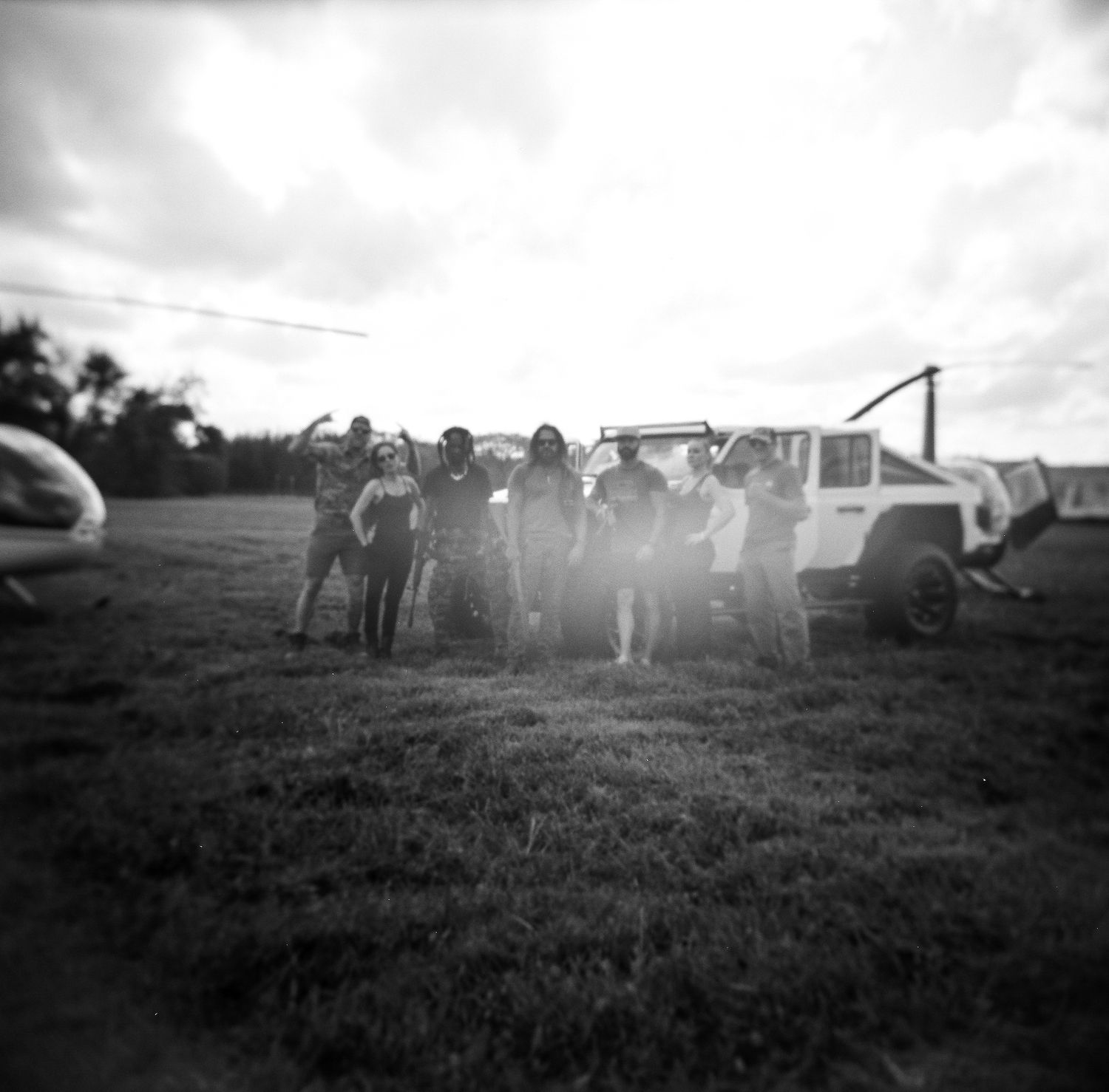 A group poses between a helicopter and white SUV on a sunlit grass field in black and white.
