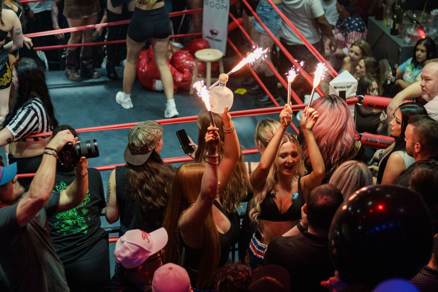 Excited women celebrate with sparklers and drinks ringside at a festive boxing entertainment event.