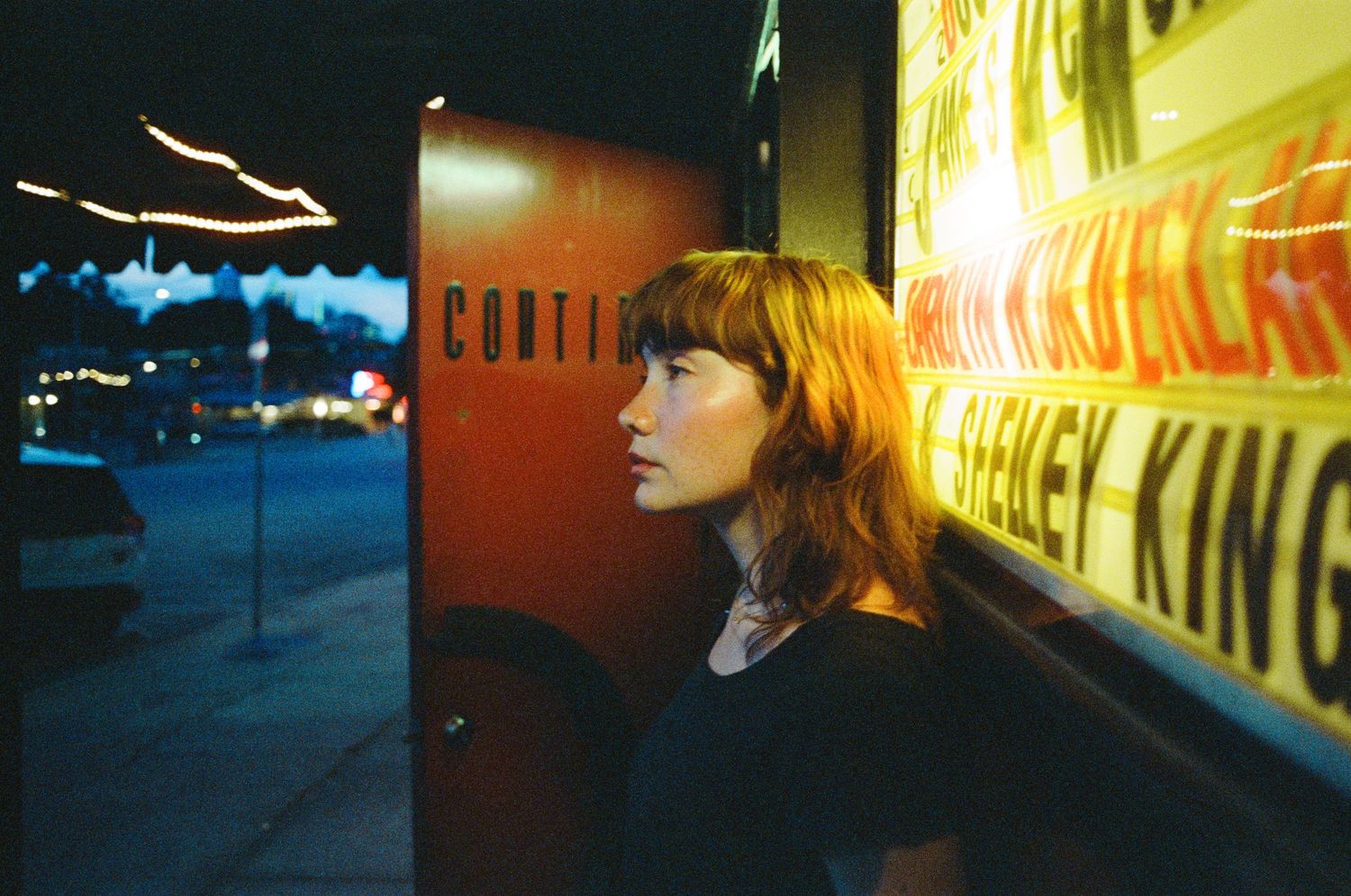 Woman with auburn hair gazes at illuminated movie theater signage at night.