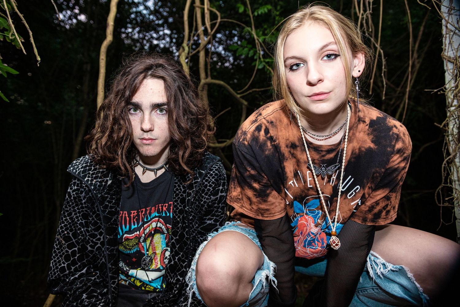 Two young teens in rock band tees pose together in a wooded outdoor setting.