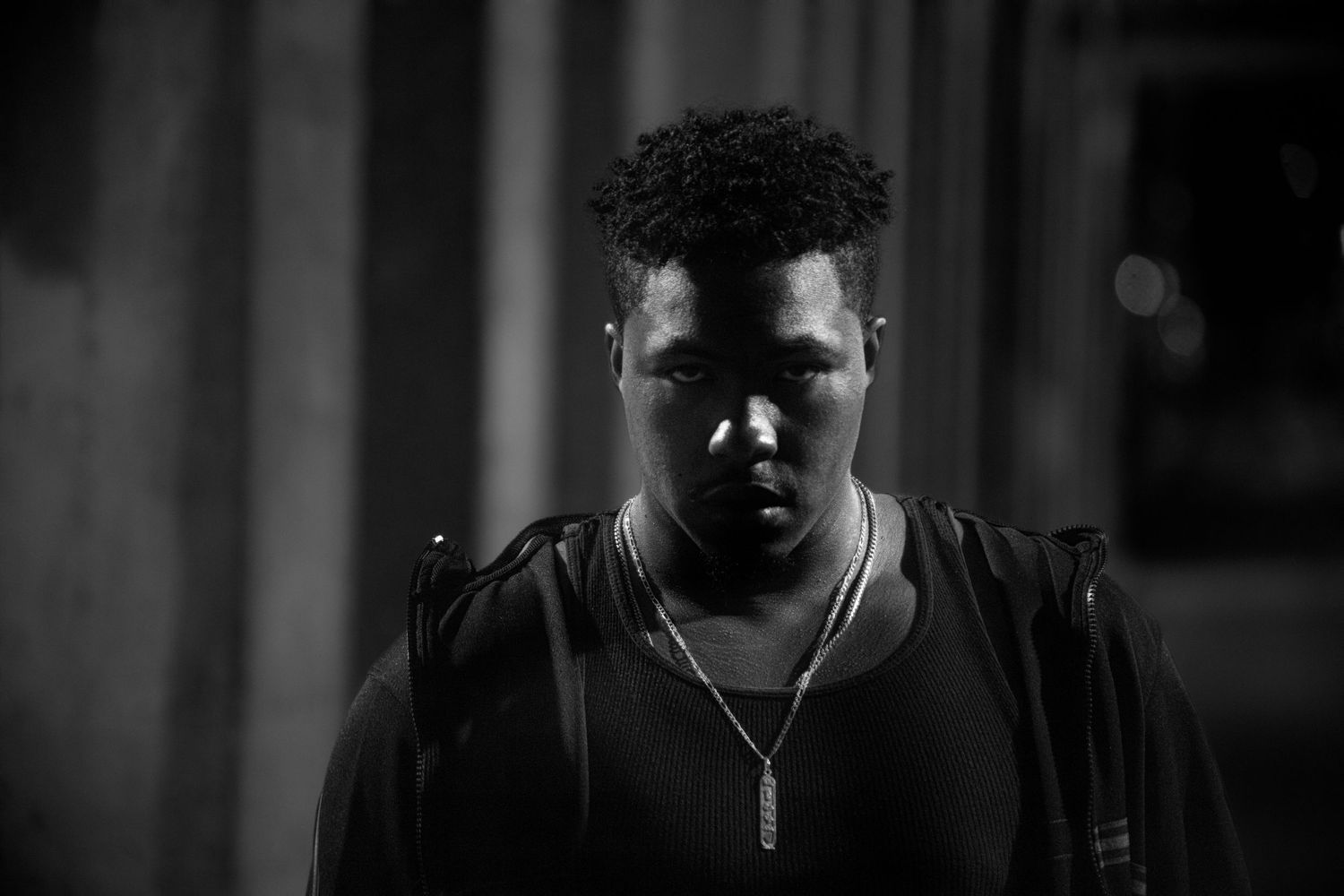 Black and white portrait of a young man wearing a chain necklace, looking down in dramatic lighting.