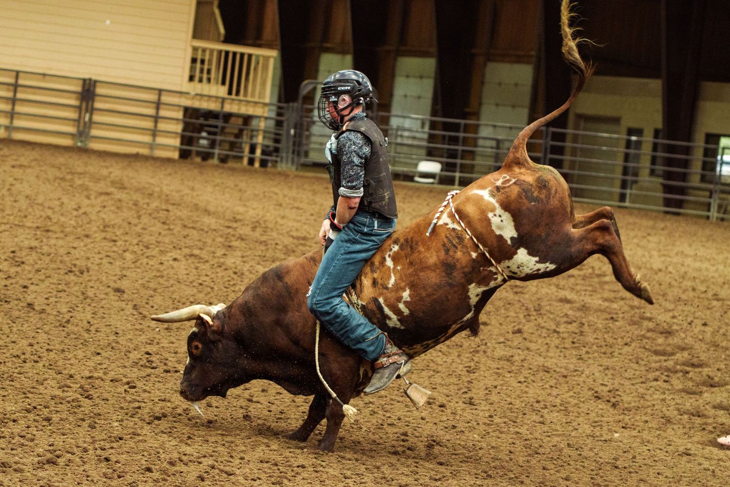 A bull rider in helmet and vest competes on a bucking brown and white bull in an indoor rodeo arena.