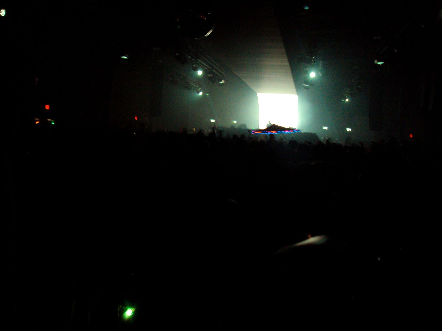 Dark concert venue with bright stage lights illuminating a large crowd at a live music event.