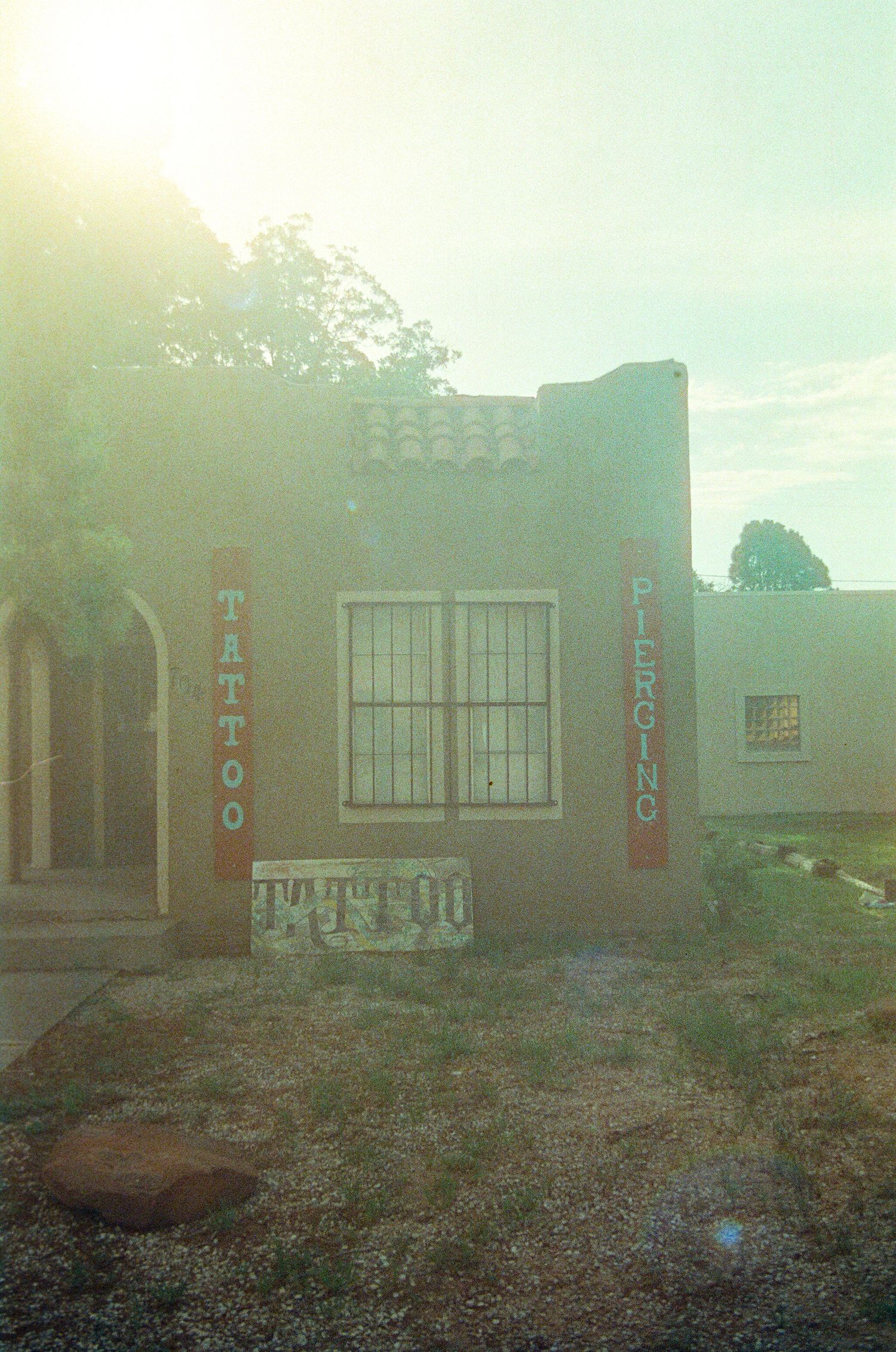 Vintage photo of a tan stucco tattoo and piercing shop with barred windows and signage in a desert setting.