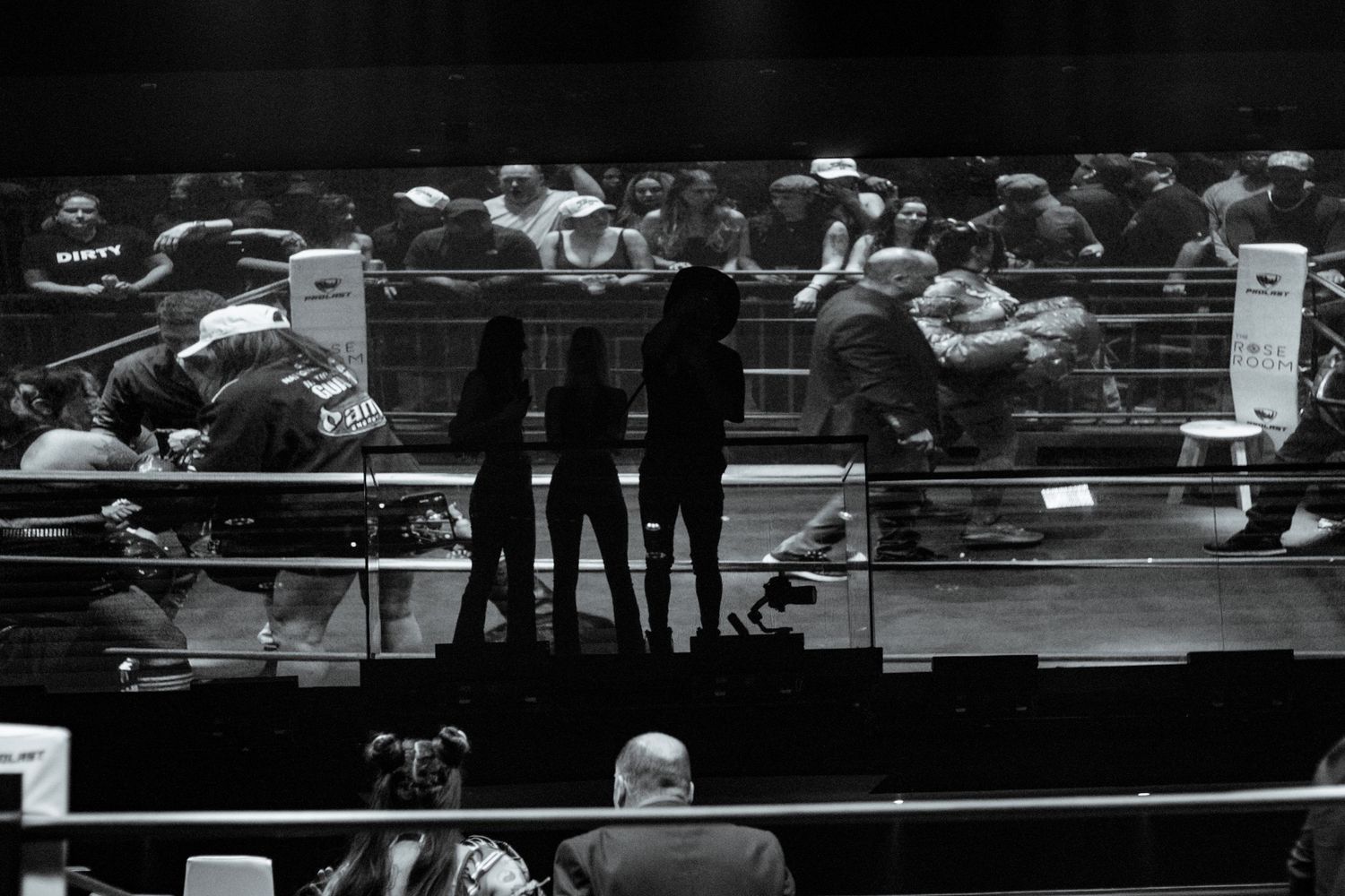 Black and white silhouette of three people standing ringside at a crowded indoor boxing event.