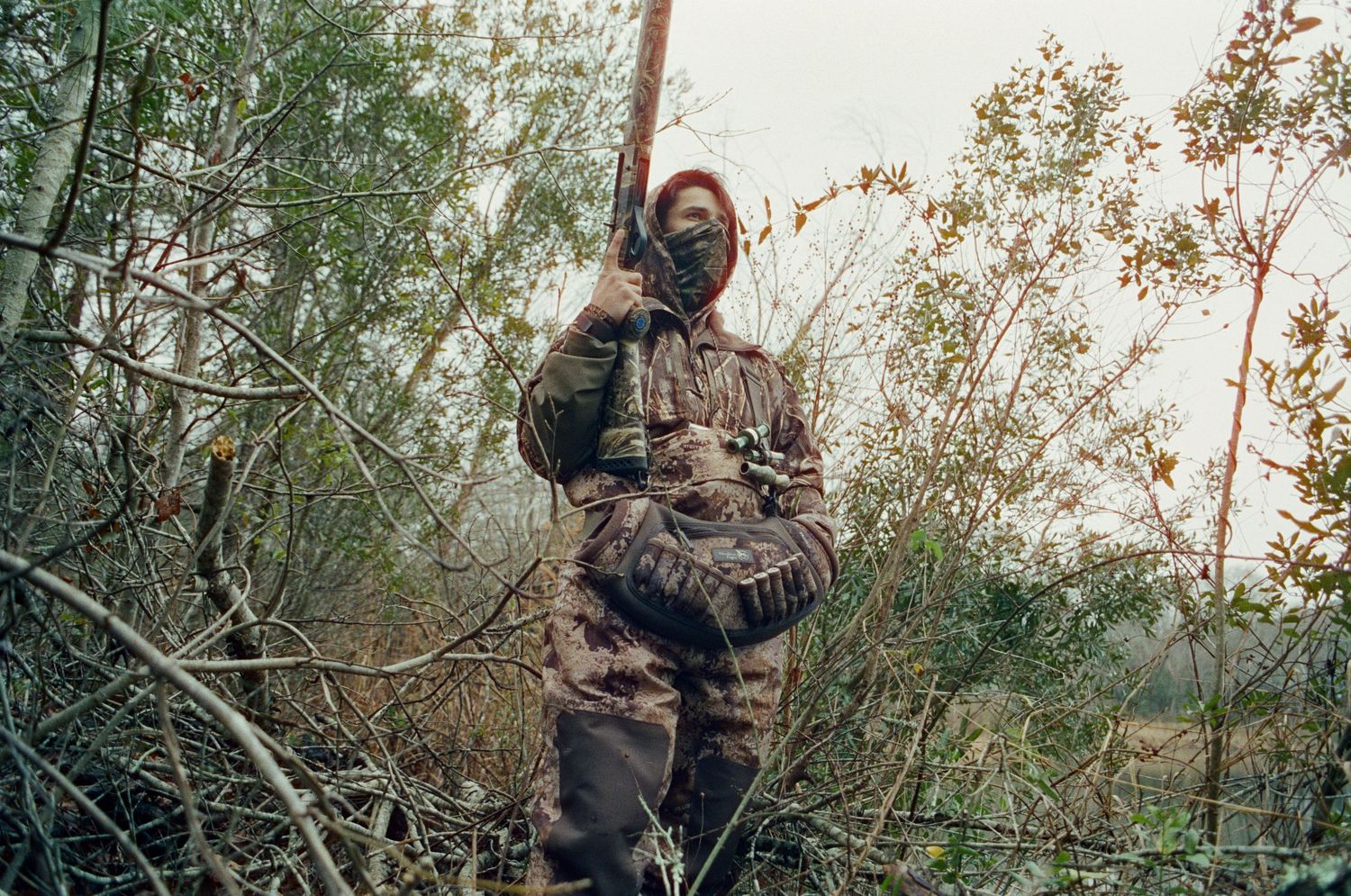 Hunter in full camouflage gear and face mask stands holding a rifle among dense brush.