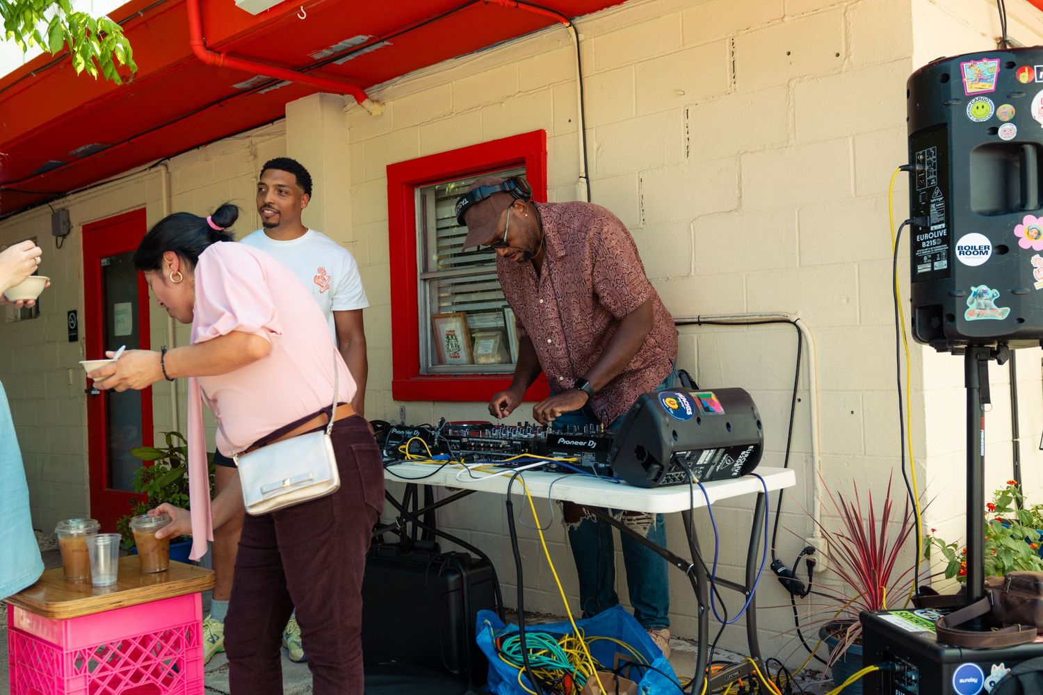 DJ and two men set up music equipment outside a red building at an outdoor event.