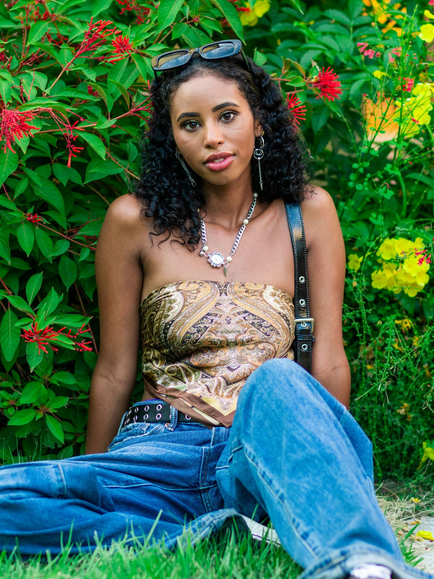 Stylish Black woman in paisley top and wide-leg jeans sits on grass surrounded by vibrant tropical flowers.