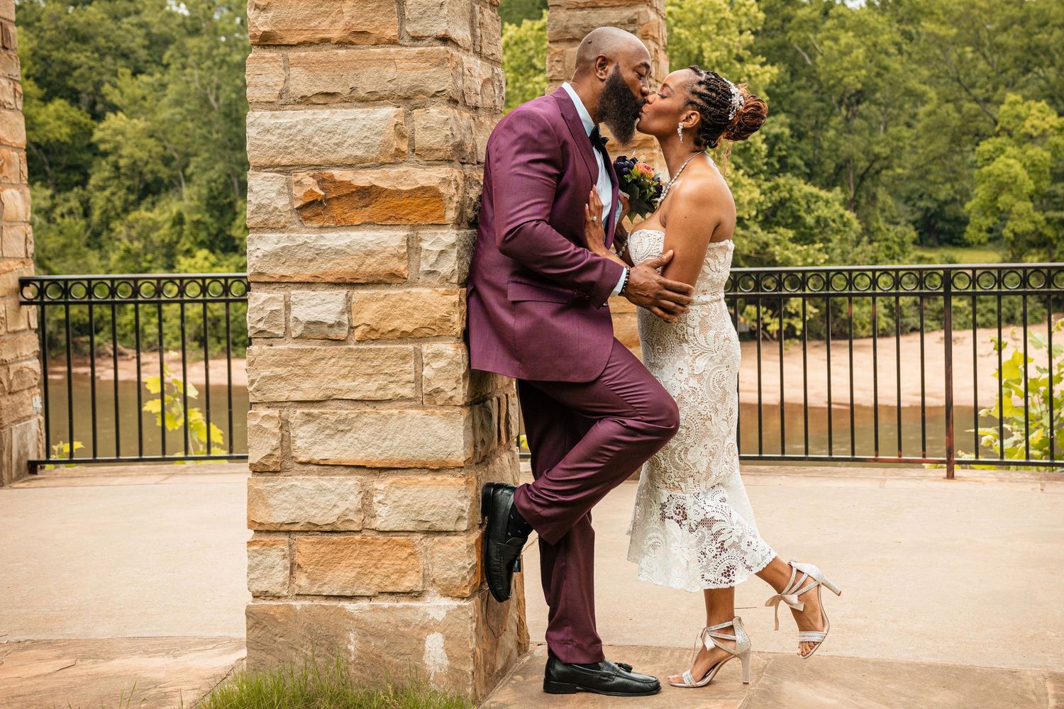 Couple sharing a kiss against a stone pillar, groom in burgundy suit and bride in fitted lace wedding dress.