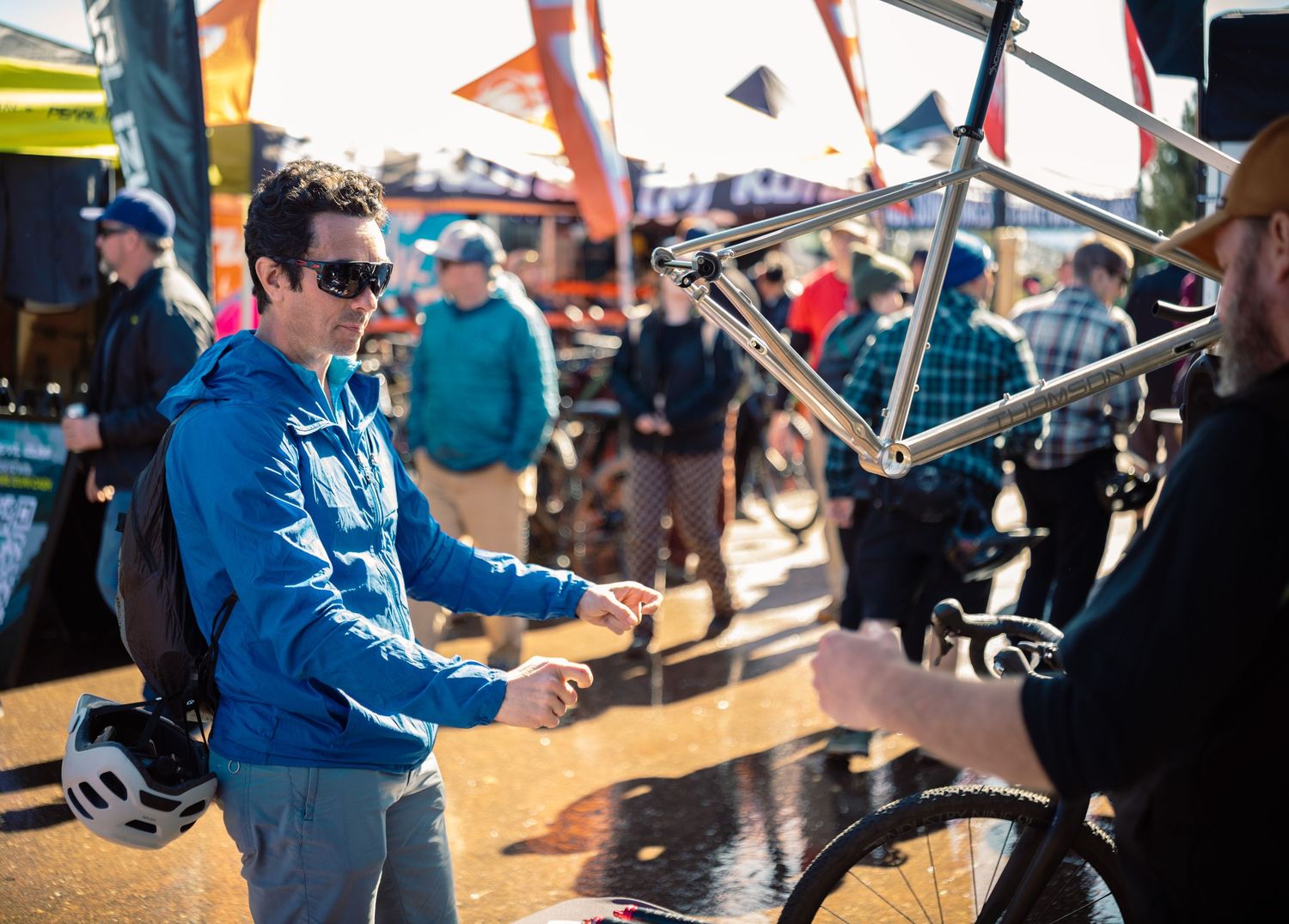 Man in blue jacket examines a titanium bicycle frame at an outdoor cycling trade show event.