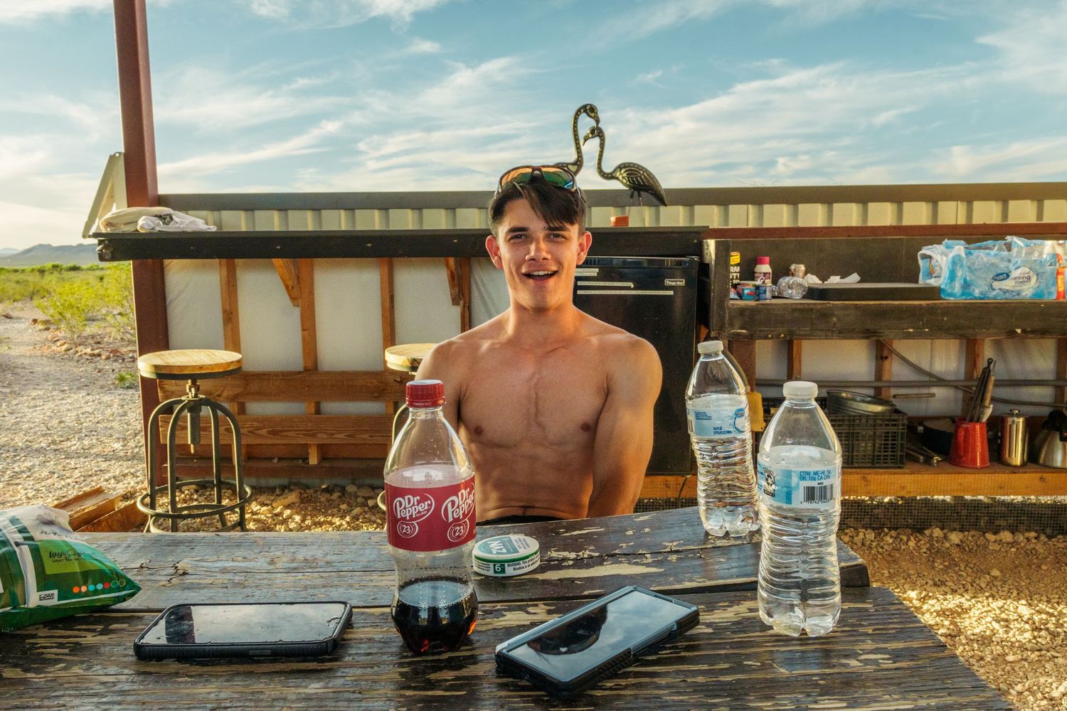 Shirtless young man sitting at outdoor picnic table with soda bottles and water in a rural setting.