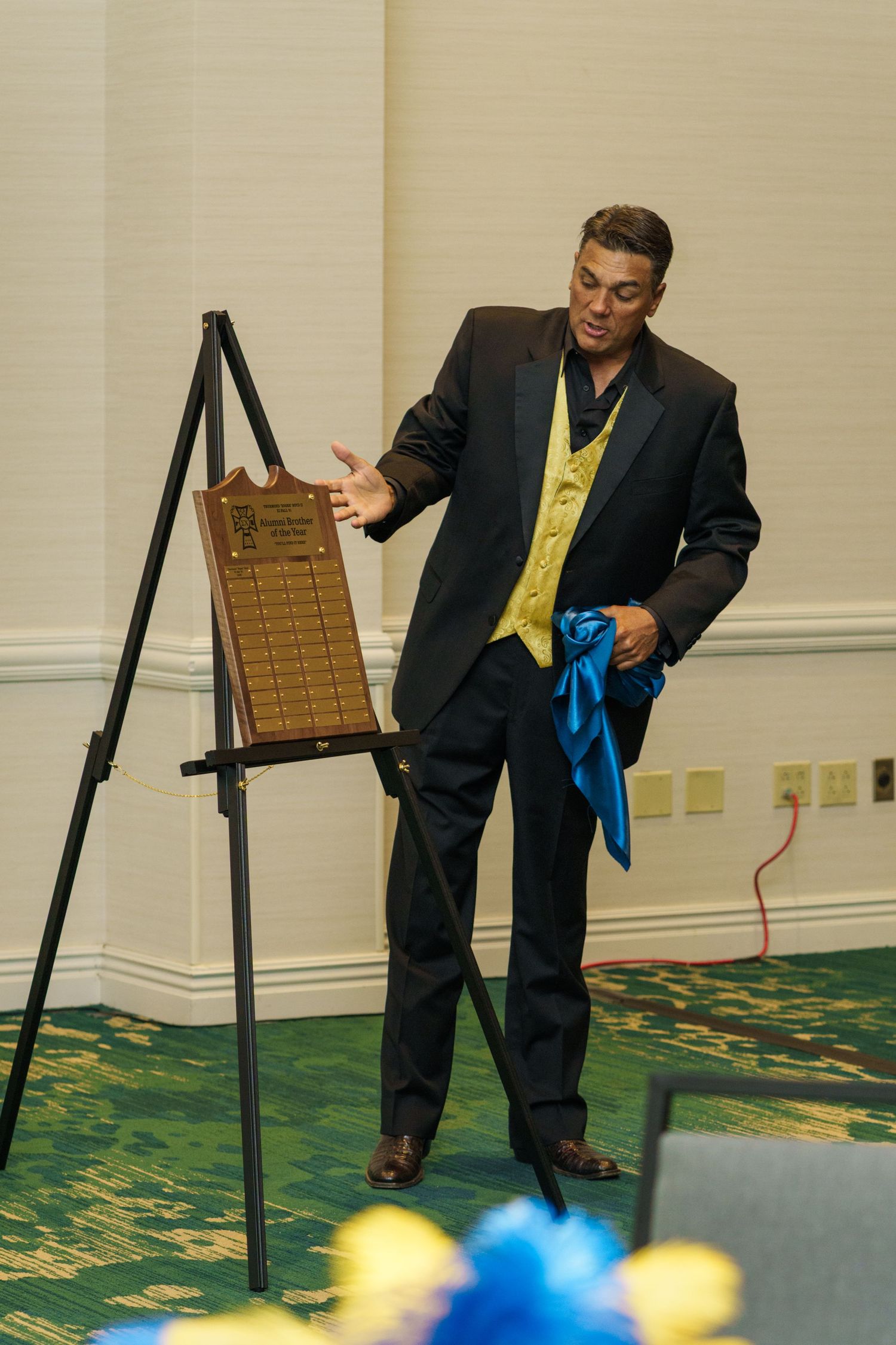 Man in black suit and gold vest gesturing toward an award plaque on an easel at a formal banquet event.