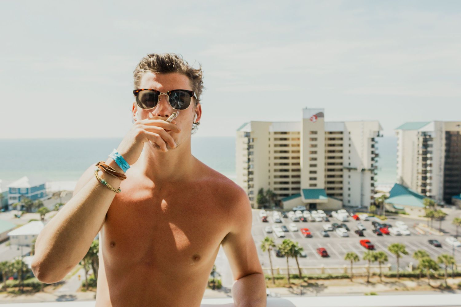 Shirtless young man wearing sunglasses on a rooftop with ocean and beachfront hotels in background.