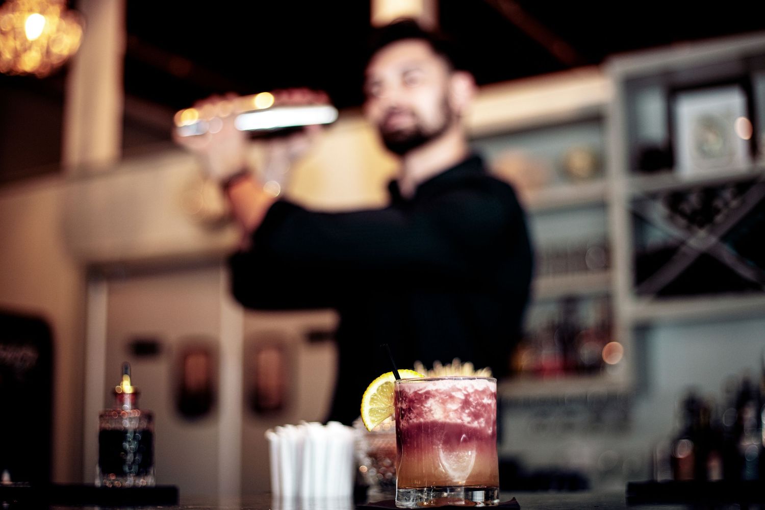 A bartender prepares a craft cocktail behind a vibrant garnished drink at a dimly lit bar.