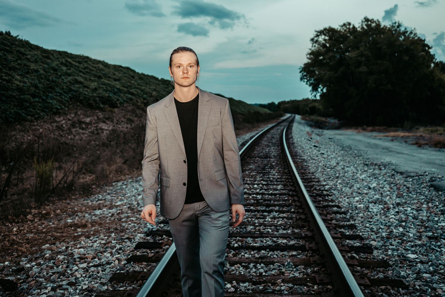 Young man in a grey blazer stands confidently on railroad tracks at twilight.