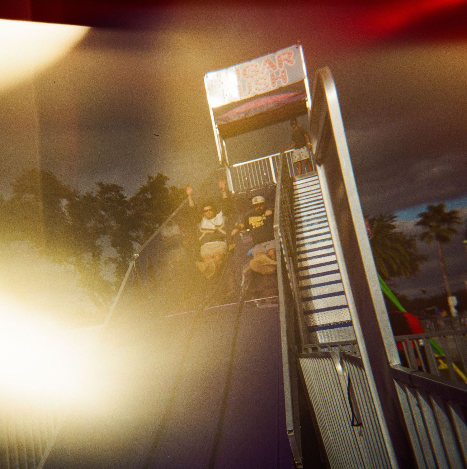 People ride a steep carnival slide attraction at an outdoor fair under a dramatic sunset sky.