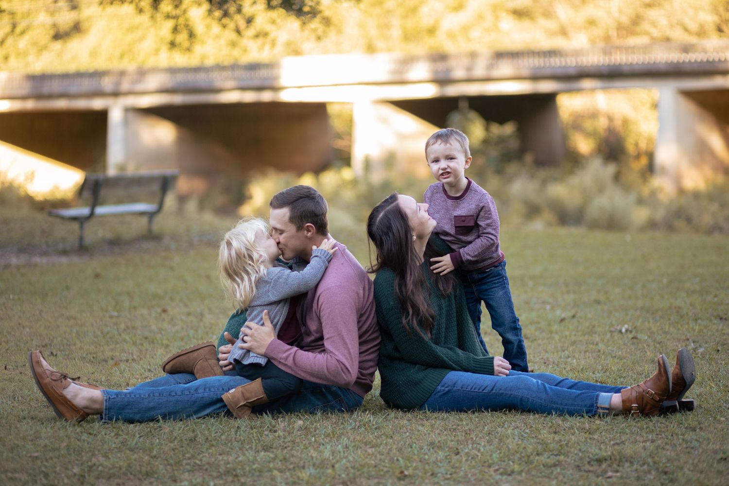 Family of five sitting on grass outdoors, parents back-to-back with playful children climbing on them.