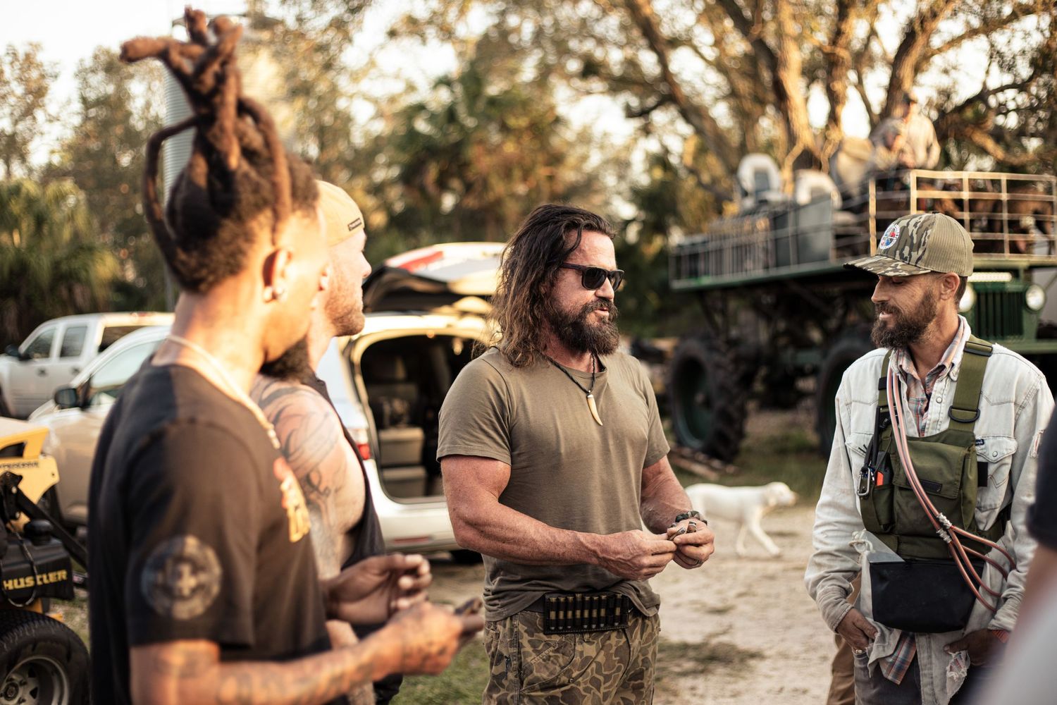 Three men in tactical outdoor gear having a conversation near off-road vehicles in a wooded setting.