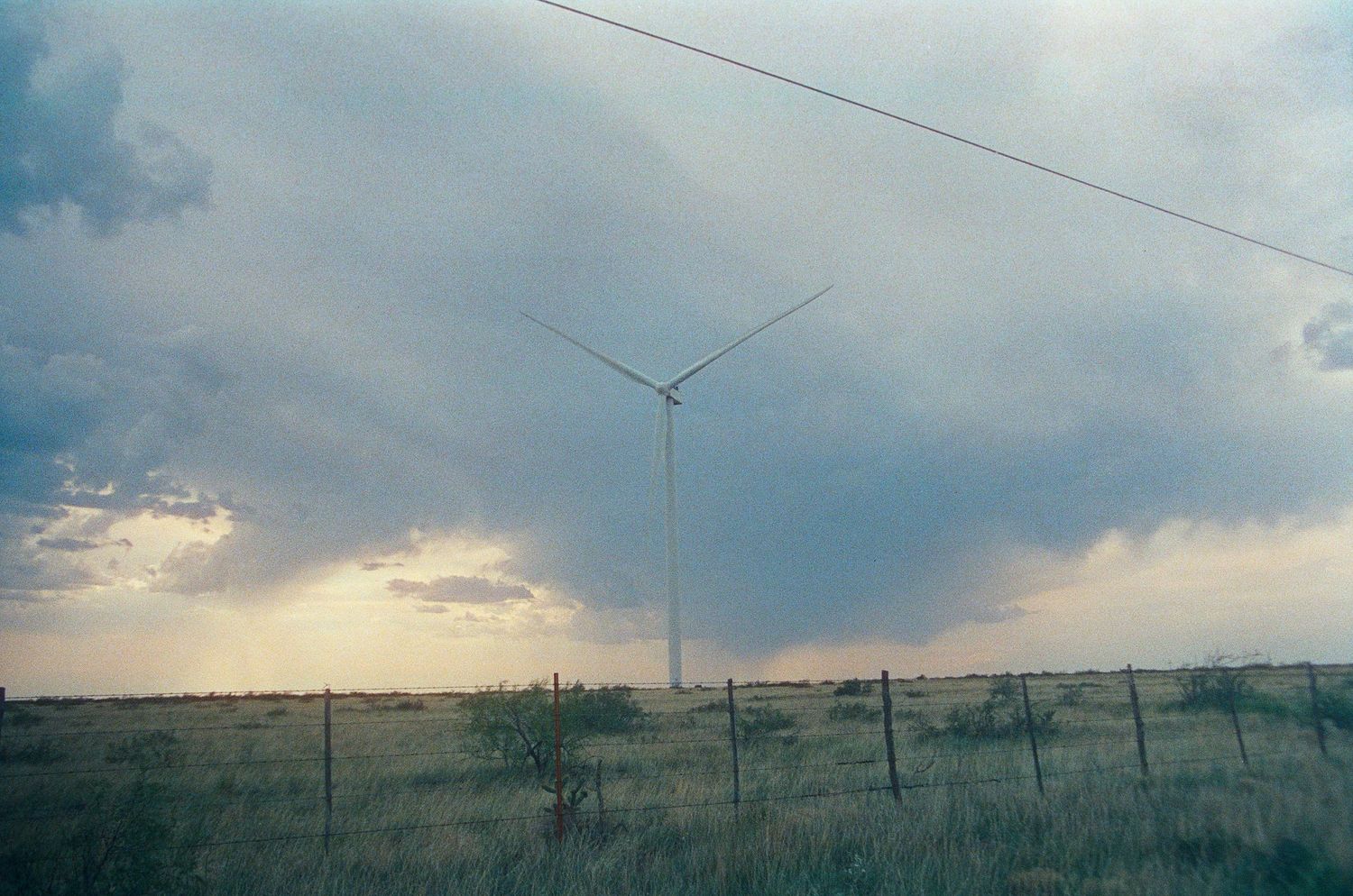 Wind turbine standing tall in a stormy field with dramatic cloudy skies and a rustic fence in the foreground.