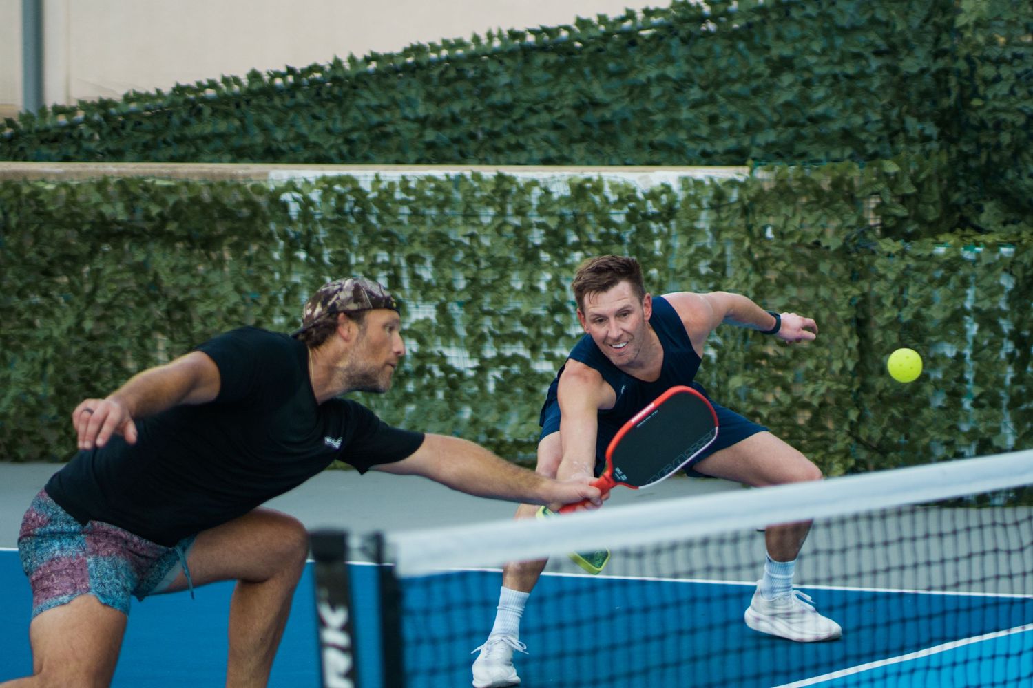 Two players compete in a doubles pickleball match, reaching for the ball near the net on a blue court.